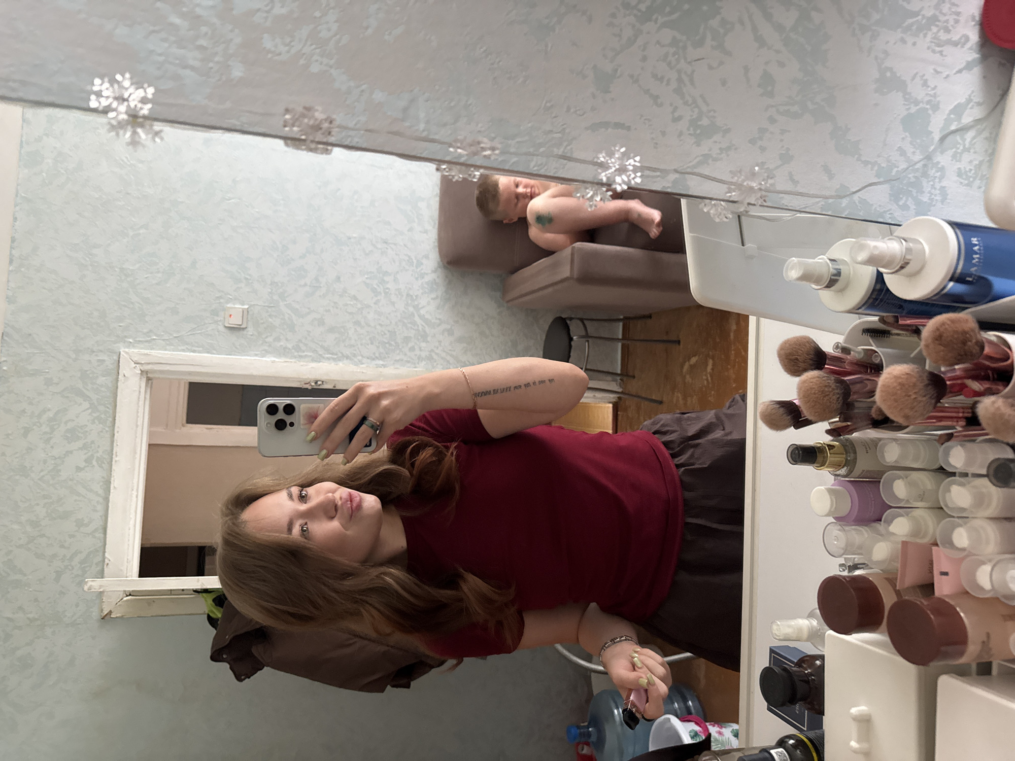 The image is a selfie taken in what appears to be a bedroom or dressing area. The photographer, a young woman with light brown wavy hair, is wearing a maroon t-shirt and dark brown shorts. She is holding a smartphone in her right hand and has her lips pursed, as if blowing a kiss. A tattoo is visible on her left forearm. Her left hand is holding a pink object, possibly a makeup item, and she is wearing bracelets on both wrists and rings on her fingers.
In the background, reflected in a mirror, is a young boy sitting on a brown armchair. He is naked from the waist up and has a green mark on his left thigh. The room has light blue patterned wallpaper. To the left of the photographer, a large water dispenser and a small stool are visible. In front of the photographer, on a vanity, are various makeup products, including brushes, bottles, and containers. There are also snowflake-shaped decorations on the mirror.
The setting is likely a residential interior. The mention of Chaikovsky, Russia, suggests this is the city where the image was taken, though the image itself does not contain explicit geographical markers. The overall impression is of a casual, personal moment captured by the photographer.