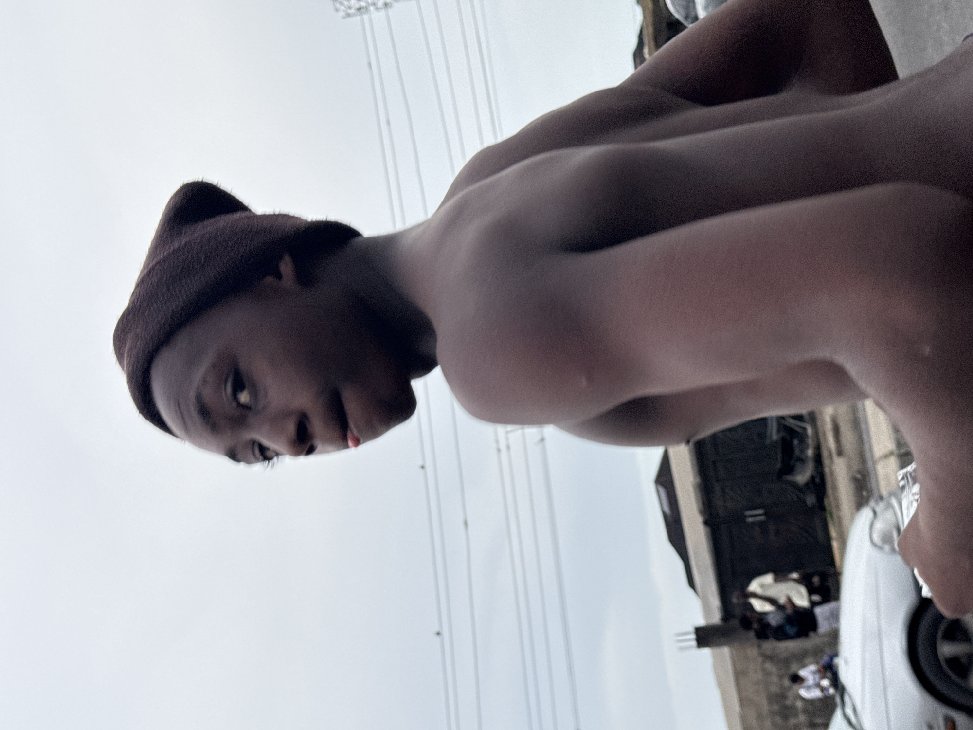 A close-up shot of a shirtless Black male wearing a brown beanie, facing towards the left. The subject has a visible car behind him with other indistinct people. Overhead electrical wires are visible in the sky. The location is identified as Ado, Nigeria.
