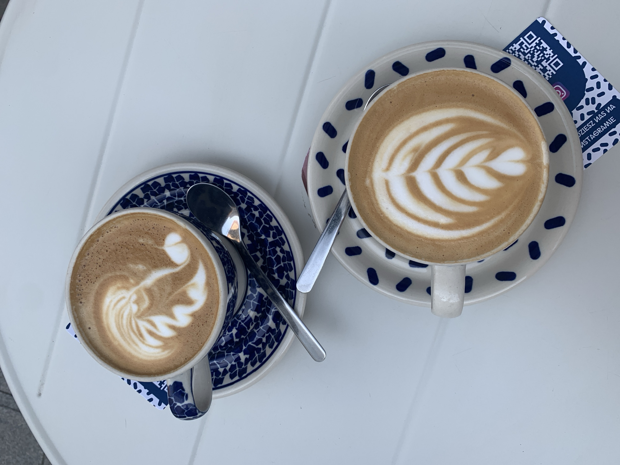 Two cups of coffee with latte art sit on saucers on a white table. Both cups and saucers have a blue and white patterned design. A spoon rests on each saucer, partially submerged in the coffee in the upper cup. The coffee in the upper cup has latte art that resembles a bird or a feather. The coffee in the lower cup has latte art that resembles a leaf. A small card with a QR code and Instagram logo is partially visible beneath the lower saucer. The table appears to be outdoors, as suggested by the light and shadow. Based on the presence of the QR code with Polish text ("Odwiedź nas na Instagramie" which translates to "Visit us on Instagram") and the distinct blue and white patterned pottery, the image is likely from a cafe in Poznań, Poland, known for its traditional patterns like the "Średzka" pattern seen on the cups and saucers.