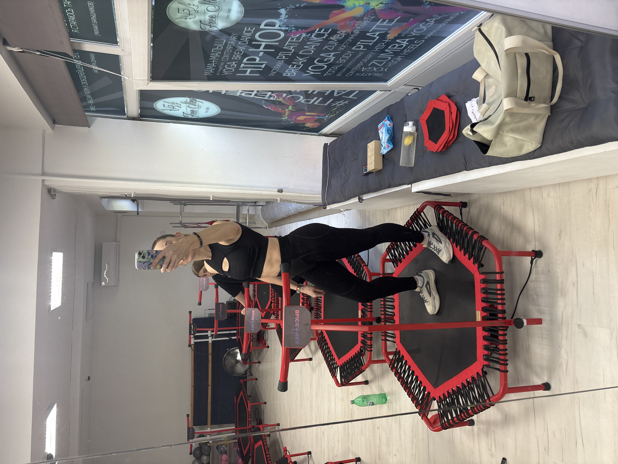 The image shows a woman taking a selfie in a fitness studio. She is wearing black workout clothes and is standing on a red mini-trampoline. In the background, there are more mini-trampolines, exercise balls, and a ballet barre. The studio appears to be well-lit and clean. A water bottle is visible on the floor next to the trampoline. The woman is holding a cell phone and taking a picture of herself in the mirror. The studio has large windows with posters advertising fitness classes such as yoga, break dance, and hip-hop. The studio is located in an urban environment, but the specific city and country are not identifiable from the image.