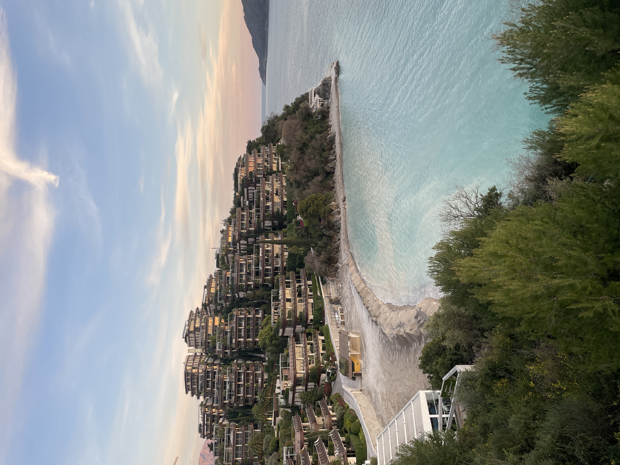The image displays a panoramic view of the Dukley Gardens resort complex in Budva, Montenegro, captured during late afternoon or early evening under a clear sky. The scene is dominated by numerous modern, terraced buildings with light-colored facades, dark frames, and extensive balconies, meticulously integrated into a verdant hillside. These structures are interspaced with lush green vegetation, including tall cypress trees and various deciduous and evergreen trees and shrubs, some showing yellow blooms in the foreground.
Below the hillside complex, a wide, crescent-shaped beach of light-colored sand or fine pebbles stretches along the shore, exhibiting linear patterns suggestive of recent grooming. A small, dark-roofed structure is visible on the beach. To the lower left, partially obscured by trees, a white, modern building with a flat roof and outdoor seating overlooks the beach. The calm waters of the bay are a vivid light turquoise-blue, with subtle ripples on the surface.
At the right end of the main beach, rocky formations jut into the water, hosting a small, multi-level white building situated directly at the water's edge. In the distance to the right, a dark, forested landmass forms a headland or island. The sky above transitions from a clear blue overhead to soft pink and orange hues near the horizon, characteristic of sunset or sunrise, with scattered wispy white clouds. A faint construction crane arm is visible in the far upper-left background. No individuals are visible within the scene.