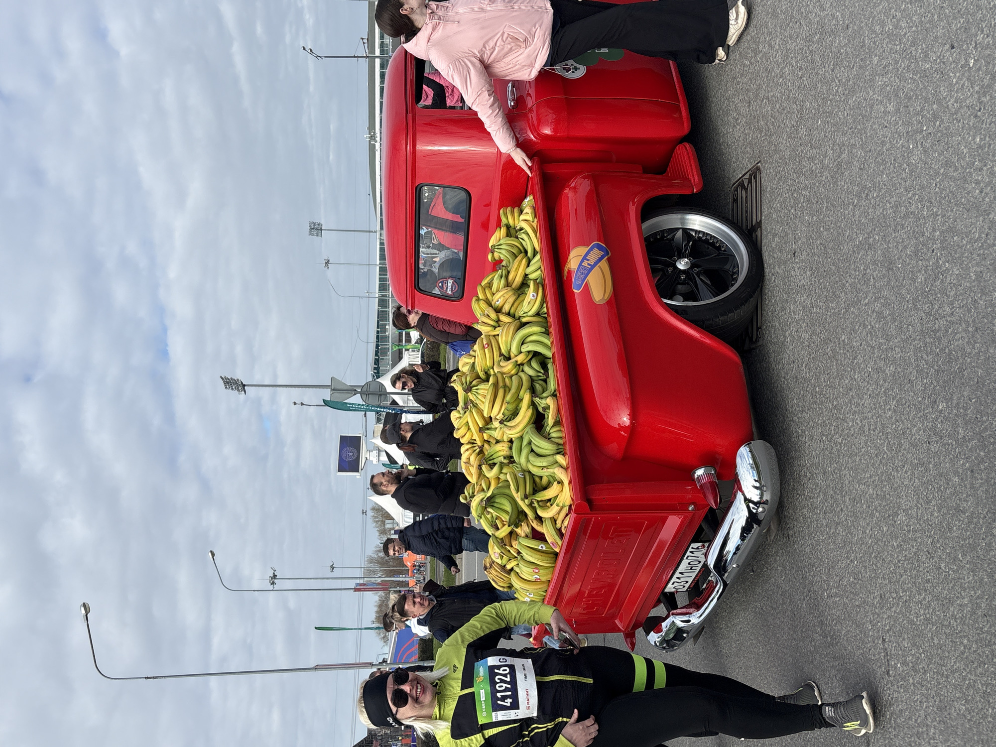 A red Chevrolet pickup truck is parked on an asphalt surface in Kazan, Russia. The bed of the truck is overflowing with bananas. A woman in a pink jacket stands on the tire and rests her hand on the truck. A second woman in black leggings and a lime green jacket lies on the ground near the truck's rear bumper. She is wearing a headband and sunglasses, and a race number "41926 G" is pinned to her jacket. Spectators stand in the background. Streetlights are visible above the crowd and a large screen displays the event's logo. A license plate with the number "0311HO716" is visible on the truck's bumper.