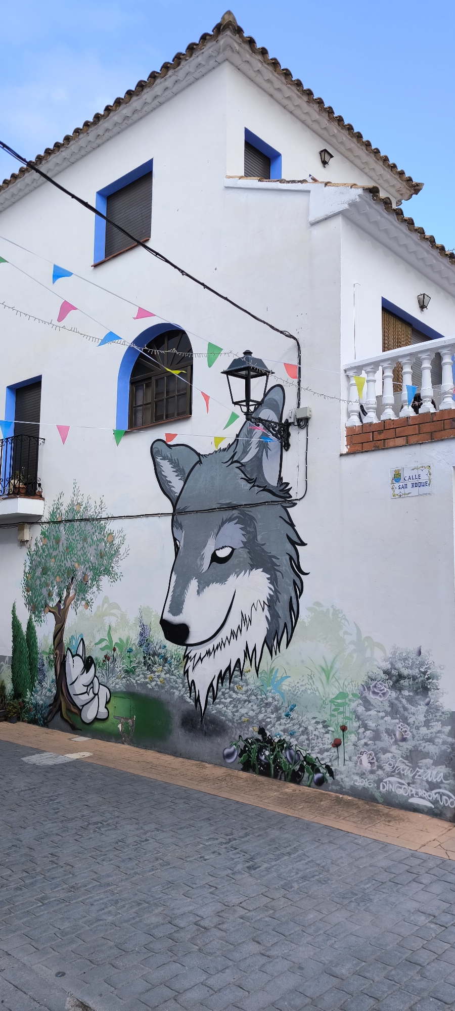 This image depicts a mural on the side of a white building in Fanzara, Spain. The mural features a large, grey wolf's head with its eyes closed and a slight smile. To the left of the wolf is a cartoonish representation of a white dog sleeping under a tree with orange fruits. The background of the mural is a colorful depiction of plants, flowers, and greenery. Decorative triangular flags are strung across the building, and a black street lamp is mounted on the wall near the wolf's head. A street sign that reads "CALLE SAN ROQUE" is visible on the right side of the building. The street in front of the building is paved with grey cobblestones. The sky above is clear and blue.
