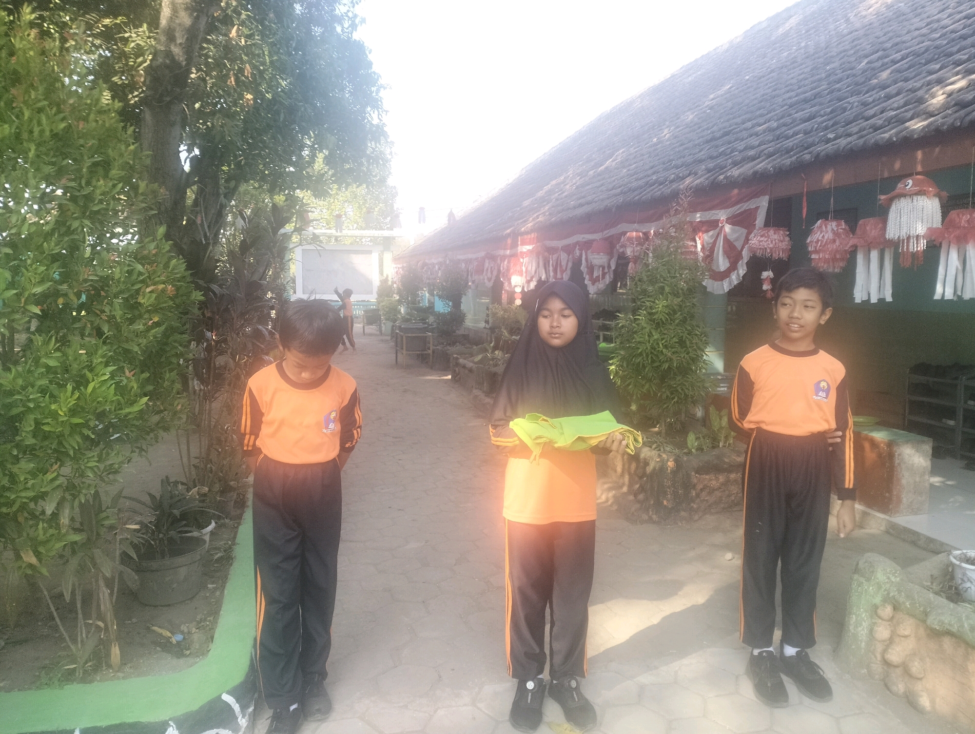 In Blabakan, Indonesia, three children, two boys and one girl, stand in a courtyard in front of a building with a tiled roof. The boy on the left, wearing an orange shirt with black sleeves and black pants with orange stripes, stands with his hands behind his back. To his right, the girl, dressed in a black hijab and an orange shirt with black sleeves and black pants with orange stripes, holds a green cloth in her hands. The boy on the right, also in an orange shirt with black sleeves and black pants with orange stripes, looks towards the right side of the frame. The courtyard is paved with stones, and lush green trees and potted plants surround the area. Red and white festive decorations hang from the building's eaves, suggesting a celebration or event. The sun is bright, casting shadows on the ground.