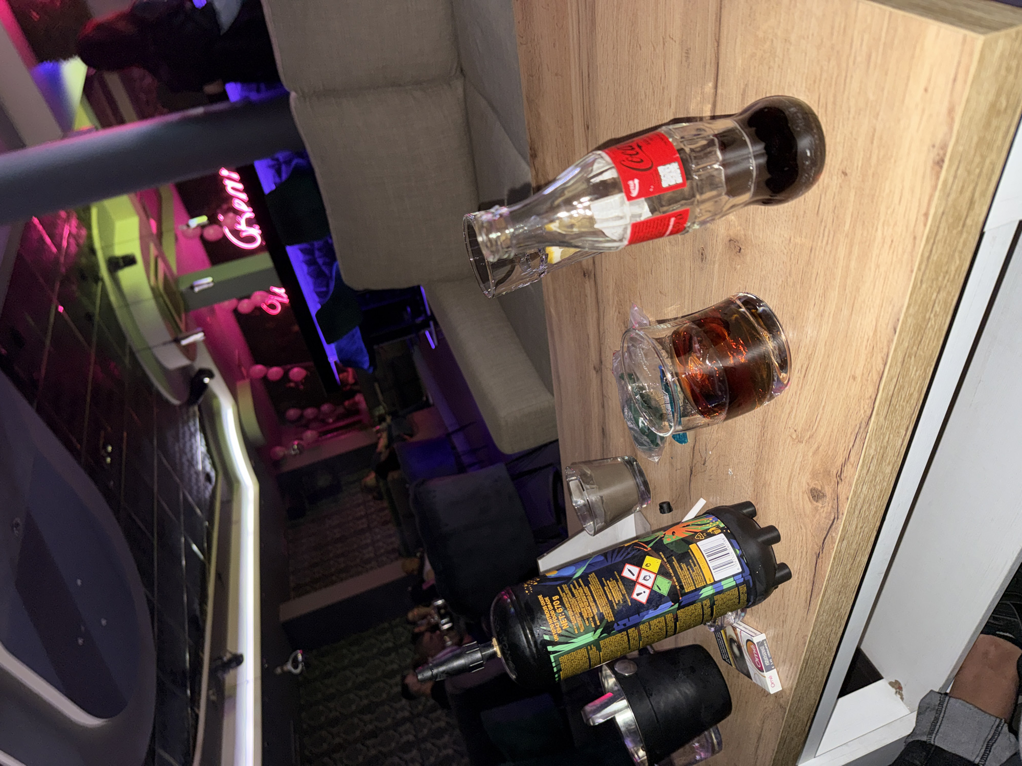 The image shows a tabletop scene with various items. On the table are an empty Coca-Cola glass bottle, a glass containing a dark liquid, an aerosol can with a colorful label, a lighter, and a pack of cigarettes. In the background, a couch is visible. The location appears to be indoors, possibly in a bar or entertainment venue, suggested by the blurred reflection of neon signs in the background. The presence of the items on the table suggests a casual social setting. The picture was taken in Karlovo, Bulgaria.