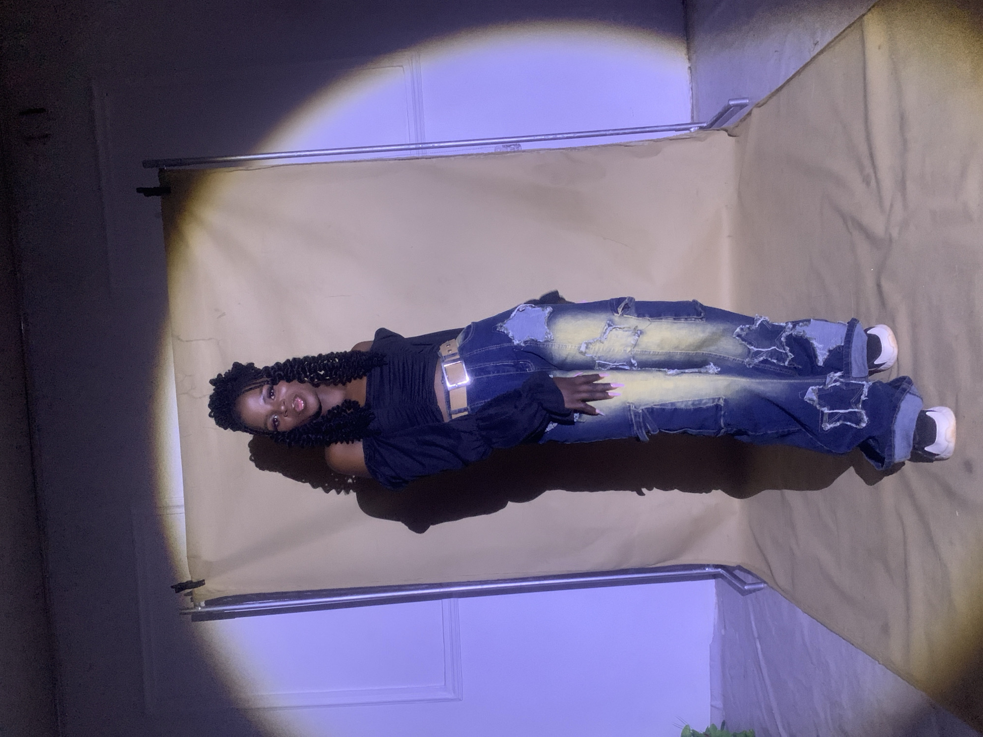 A young, dark-skinned woman with dark curly hair is lying on a light brown backdrop in a room. She is wearing a black top, ripped blue jeans with a yellow wash, and white sneakers. Her nails are pink. The backdrop is attached to a metal frame. The walls of the room are white and are bathed in a blue light. The location cannot be verified as Jos, Nigeria.
