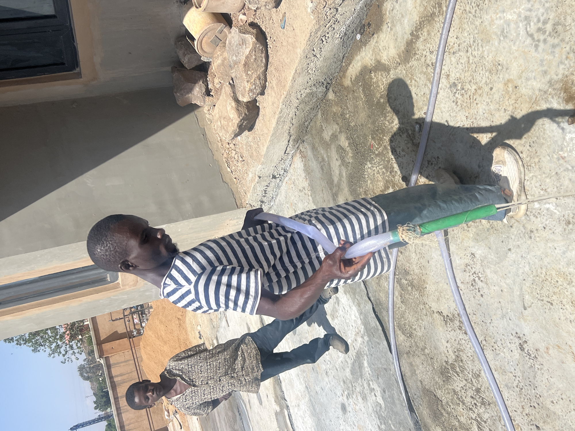 In Barakin Lerawi, Nigeria, two African men are outdoors in bright sunlight on a concrete surface next to a building. The man in the foreground wears a striped shirt and dark pants, holding a connected hose. A second man, wearing a patterned shirt and dark pants, stands further back in the scene. Near the building are rocks and possibly pottery fragments. The background includes a fence and a tower.