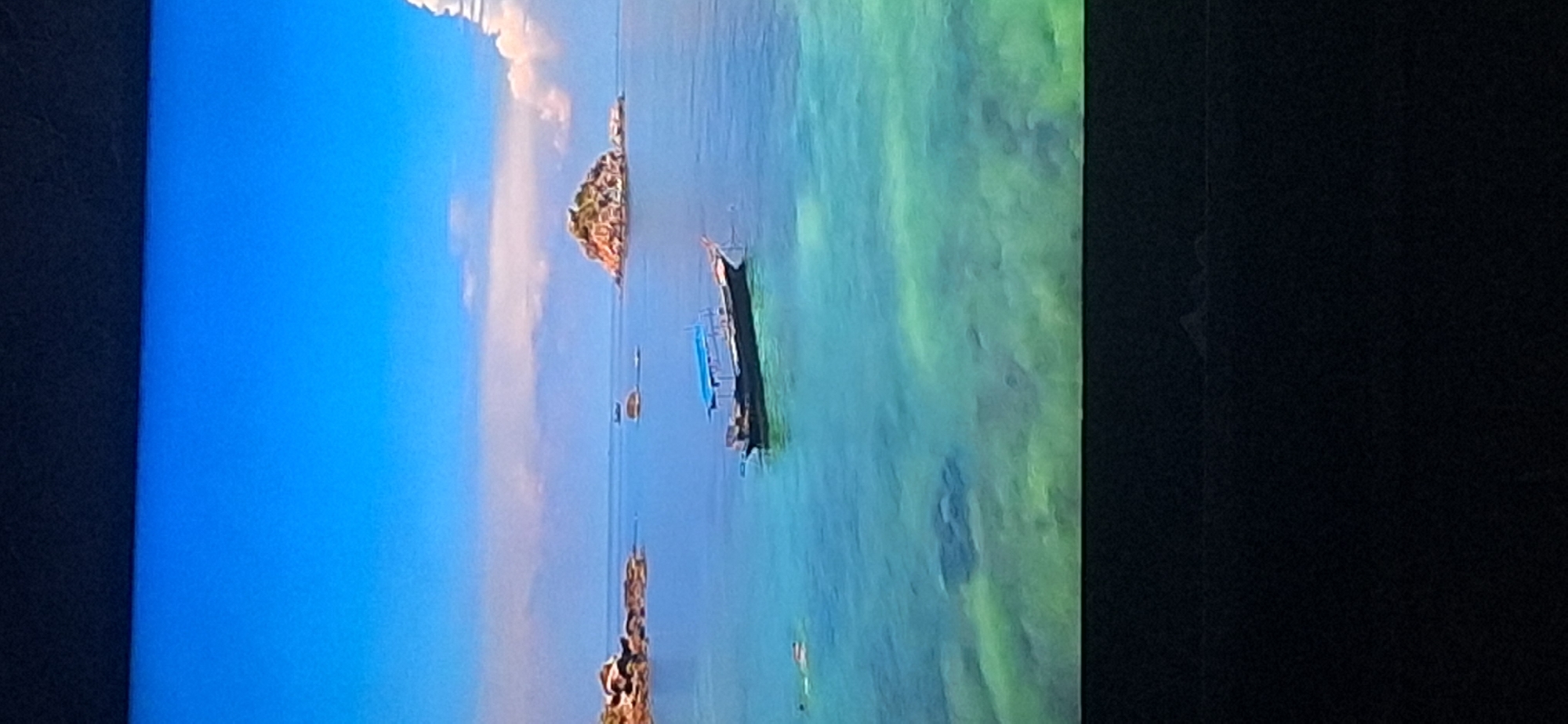 The image shows a scenic view of a tropical coastline. The sky is a clear, vibrant blue with a few scattered clouds. The ocean is calm and a striking turquoise color, with visible coral reefs or sandy bottom beneath the surface. In the foreground, a long, dark-colored boat with a blue canopy is anchored in the water. To the left of the boat, two individuals are swimming or snorkeling, indicated by their visible heads and fins. In the distance, several small, rocky islands with lush vegetation dot the horizon. The overall impression is one of a serene and picturesque tropical paradise. This image is not related to the city of Bila Tserkva, Ukraine.
