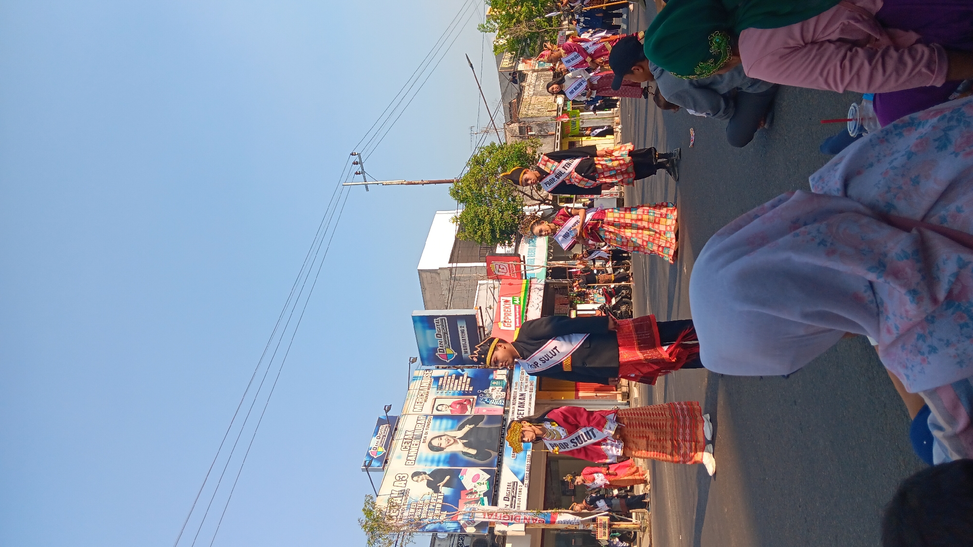 The image captures a street parade in Tanjung Anom, Indonesia. In the foreground, several people are seated on the asphalt, some wearing hijabs. The main subjects are a group of young individuals dressed in traditional Indonesian attire, participating in the parade. They are wearing ornate headpieces, vibrant sarongs, and sashes with text, likely indicating their roles or affiliations. One individual wears a black jacket over a red and gold patterned sarong, with a white sash reading "PROP. SULUT." Another person in front of him wears a red top with gold trim and a matching sarong, also with a "PROP. SULUT" sash. Behind them, other participants in similar traditional clothing are visible, along with spectators lining the street. The background shows buildings with advertisements, including one for "DANI DIGITAL PRINTING SOLUTION." Power lines crisscross the clear blue sky, and trees are visible along the street. The scene appears to be during daytime, with bright sunlight casting shadows.