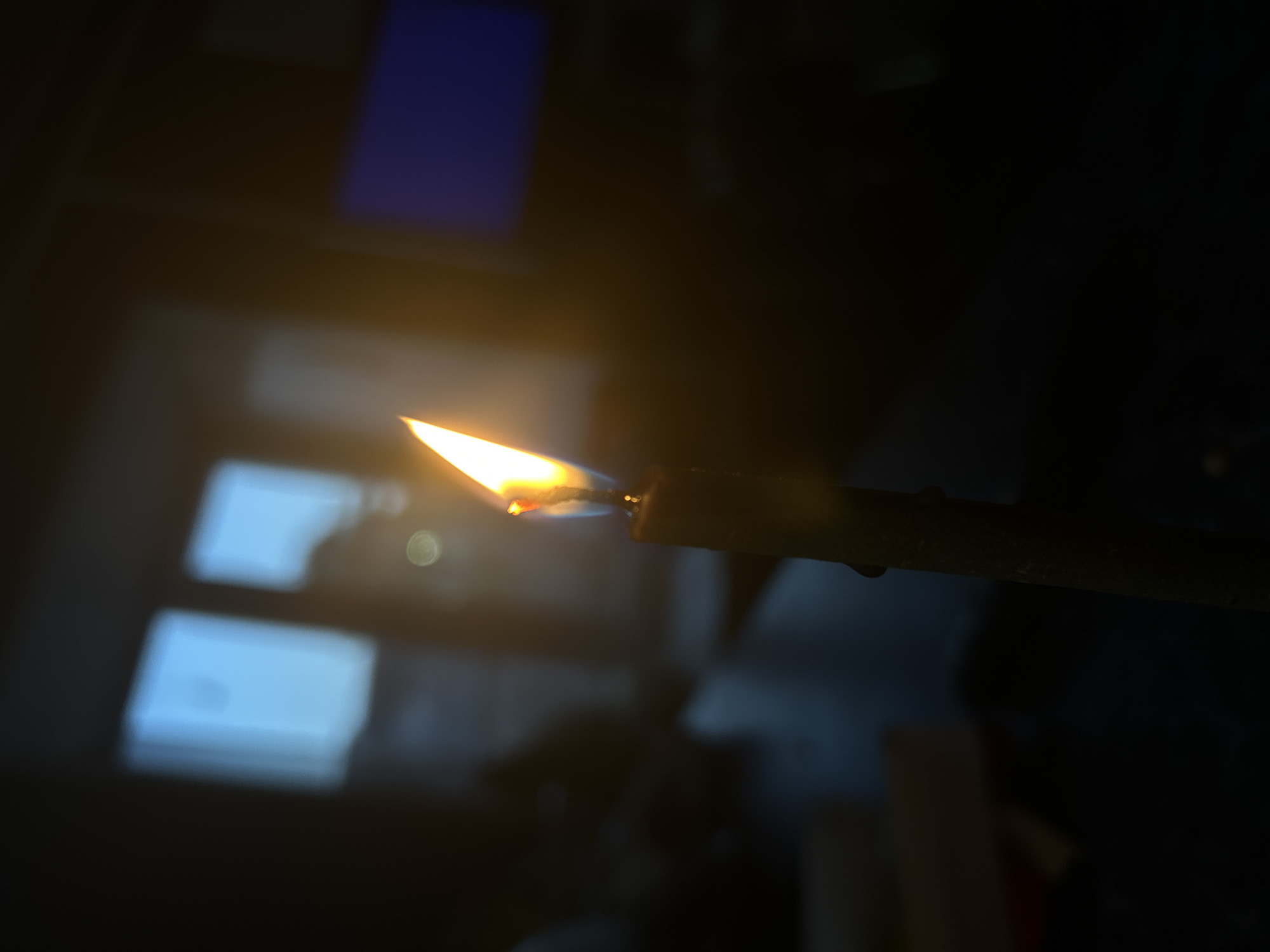 The image shows a lit candle in a dark room. The flame is the brightest object in the frame, with a yellow and orange hue. A dark, thin candle is held horizontally, with a small droplet hanging off the side. The background is blurry and indistinct, but appears to show a dark surface with some light reflections from unseen objects. There is a vague impression of some objects in the background, including rectangular shapes. There is no indication of the location being Rusalevka, Russia or any specific people.