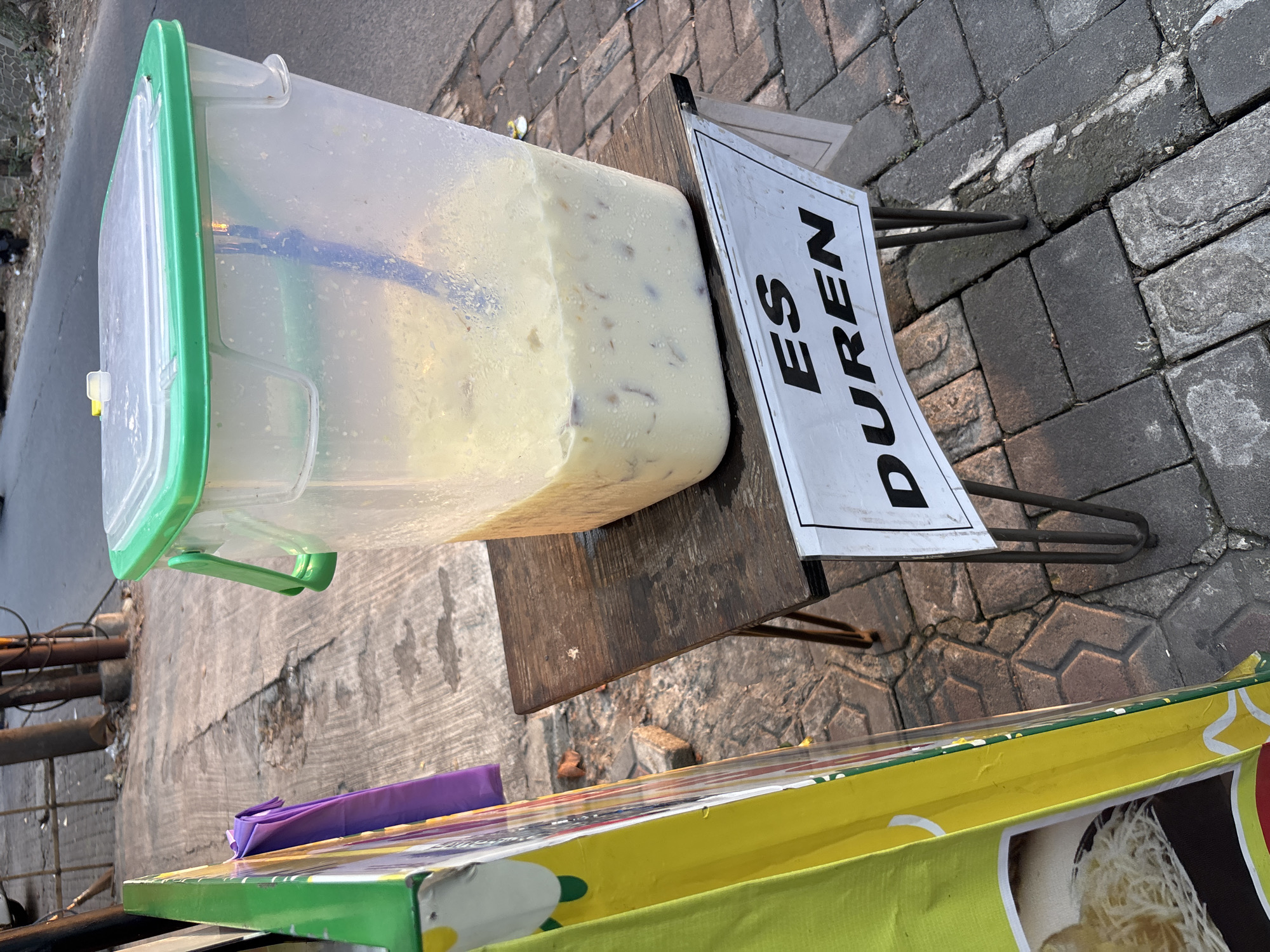 A tall, clear plastic beverage dispenser with a green lid and handle sits on a dark wooden table on a sidewalk. The dispenser contains a light-colored, milky liquid with chunks of fruit or ice. A blue ladle is inside the dispenser. A white sign with black text reading "ES DUREN" is attached to the front of the table, with the letters "ES" on the top line and "DUREN" on the bottom. To the left of the table is a portion of a tall, yellow and green sign, with a circular image and white text visible. A purple cloth is draped over a railing behind the dispenser. The sidewalk is paved with interlocking gray bricks. The scene appears to be outdoors, possibly a street vendor stall in Bekasi, Indonesia, with a road and some debris visible in the background. The lighting suggests it is late afternoon or early morning.