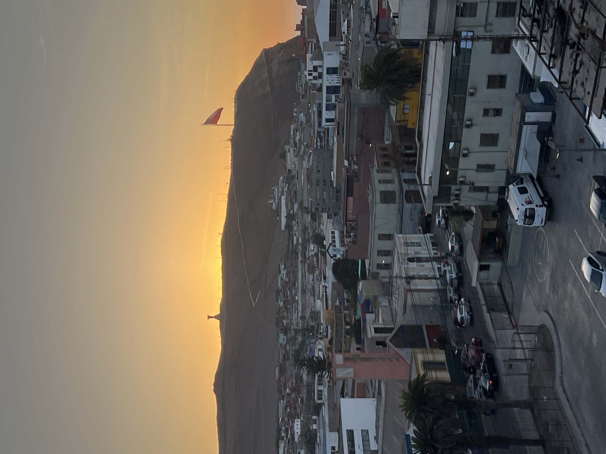 The image shows a panoramic view of Arica, Chile, during sunset. A large hill dominates the left side of the frame, topped with a statue and a large Chilean flag. The city stretches out toward the right side, filled with buildings of varying heights and colors. Streets with parked and moving cars are visible. The sky has a gradient of orange and yellow hues. A parking lot is situated in the foreground on the right side.