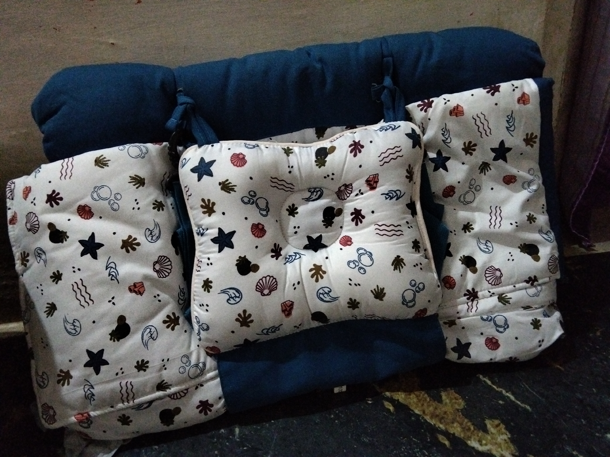 The image shows a set of three baby pillows with a white fabric cover featuring a repeated pattern of seashells, starfish, seaweed, and bubbles in various colors like navy, pink, and brown. The pillows are arranged on top of a dark blue, padded mat or cushion. Two of the pillows are rectangular, and one in the center is more rounded and has a slightly indented area, likely for a baby's head. The pillows are tied with navy blue fabric ribbons to the larger blue mat. The background is a textured wall and a dark, speckled floor. The scene appears to be indoors, possibly in a residential setting. The city of Benuaraja, Indonesia is not depicted in this image.