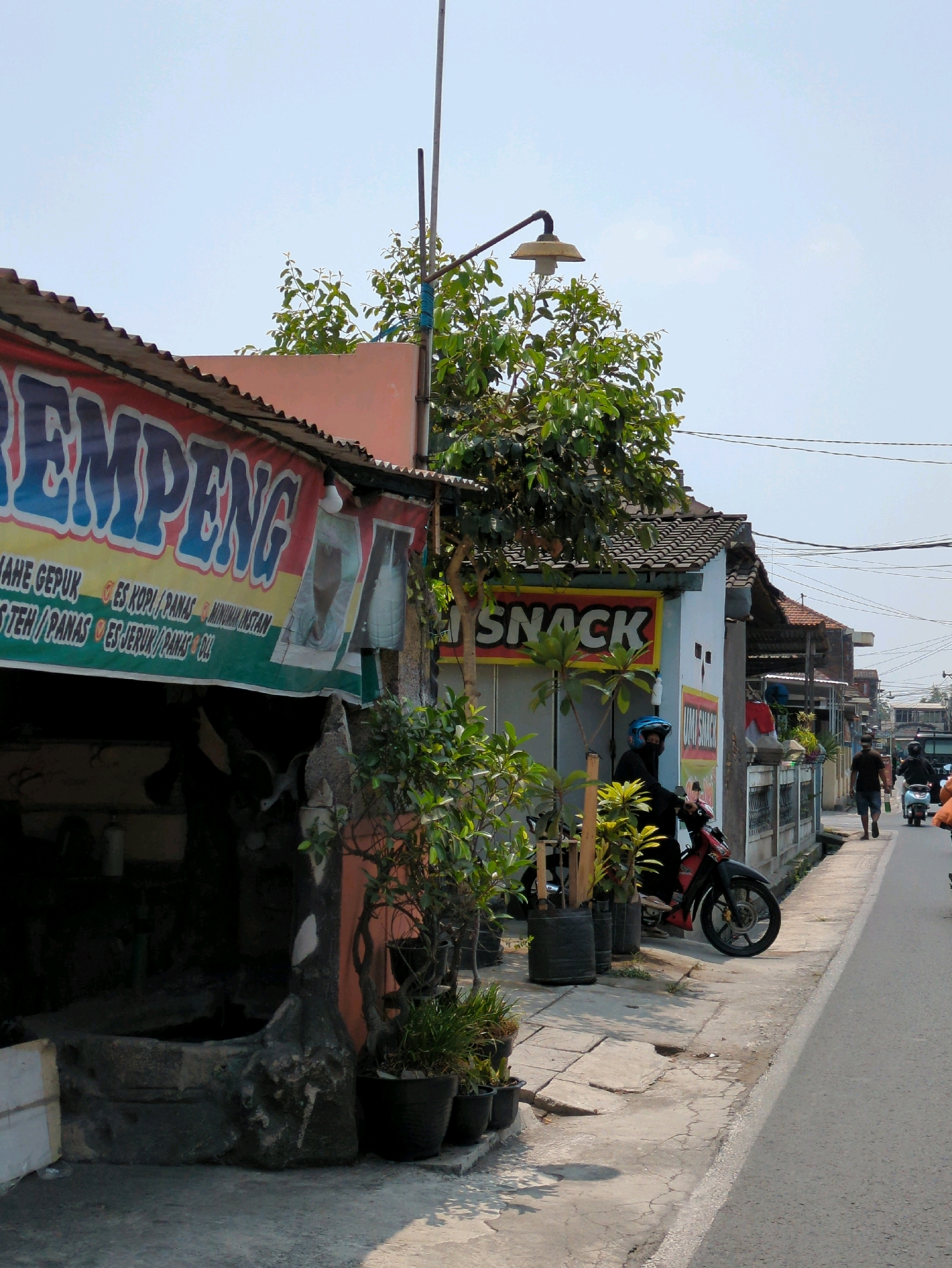 The image depicts a street scene in Gumpang, Indonesia, likely on a bright, sunny day. On the left, a building with an orange facade features a large banner with red and blue lettering that reads "REMPENG," indicating a food or drink establishment. Below this, smaller text lists menu items such as "MAHE GEPUR ES KOPI PANAS MINUMAN INSTAN" and "ISTEH PANAS ES JERUK PANAS OLL." A dark, possibly open-air area with some objects is visible beneath the banner.
Adjacent to this building, another structure with a light blue wall displays a yellow sign with red lettering that says "SNACK." In front of this, a red motorcycle is parked, and a person wearing a black outfit and a blue helmet is seated on it. Several potted plants are arranged along the sidewalk.
The sidewalk itself is made of concrete paving stones and shows signs of wear and tear. A grey stone wall with some decorative elements separates the sidewalk from a further building or area. Further down the street, two men are walking, and other motorcycles and a vehicle are visible in the distance, suggesting moderate pedestrian and vehicular traffic. Power lines are strung overhead. The overall impression is of a typical street scene in a residential or commercial area of an Indonesian town.