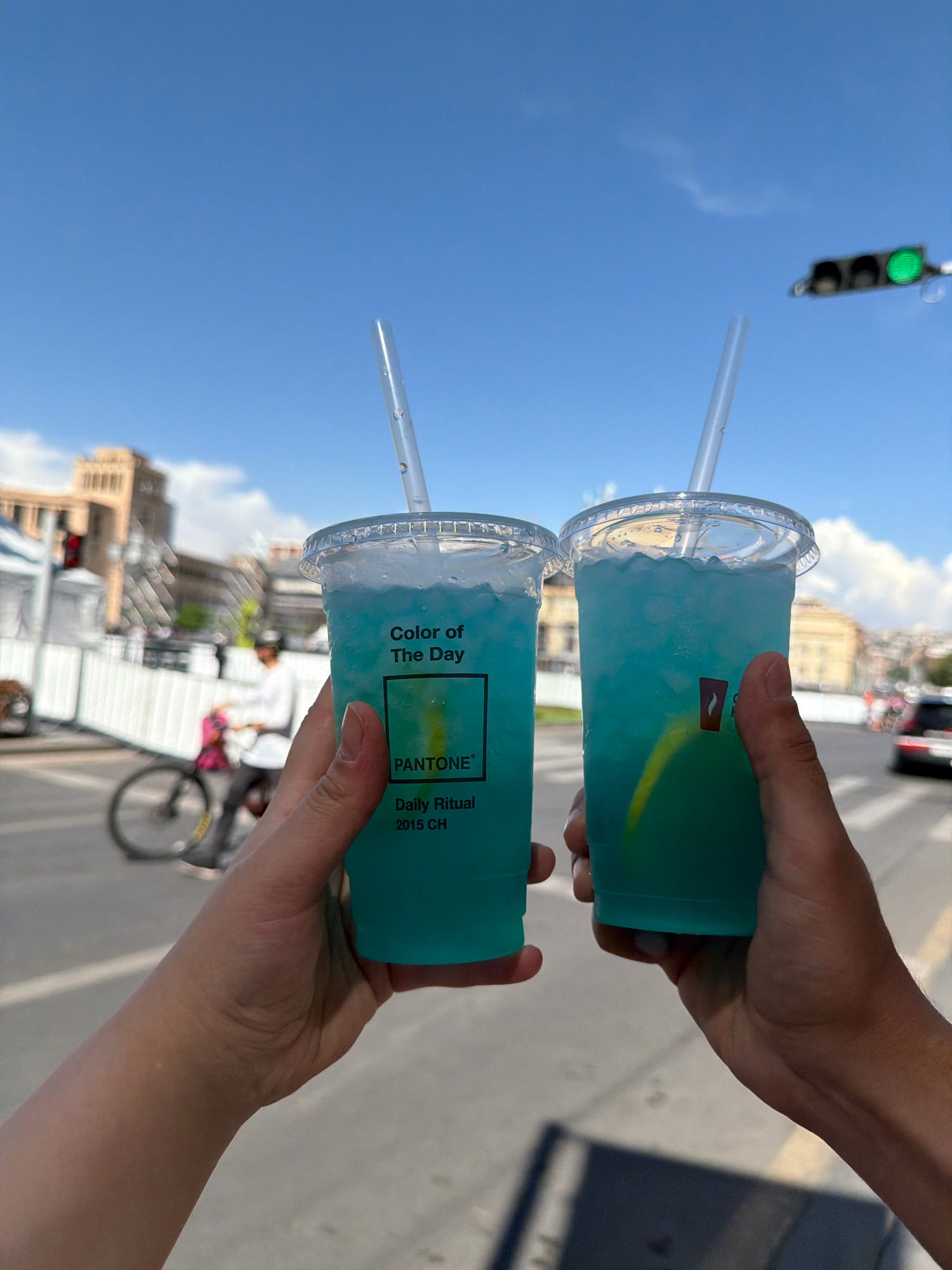 Two hands are holding up two clear plastic cups filled with blue liquid and ice, and straws. The cups have labels that say "Color of The Day," "PANTONE," and "Daily Ritual 2015 CH." The background is a city street with buildings, a traffic light with a green signal, and a person riding a bicycle. This scene is the Republic Square in Yerevan, Armenia, on a sunny day with a clear blue sky and some clouds.