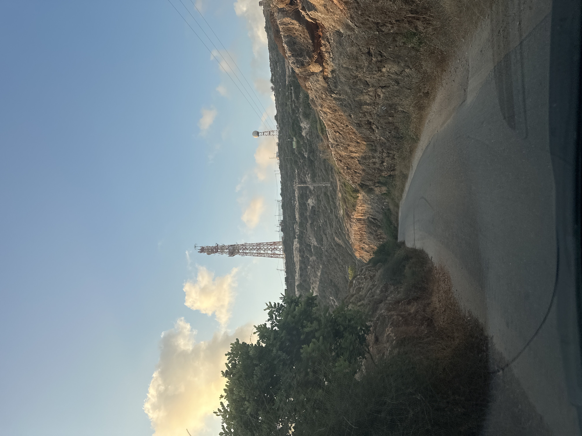 The image depicts a paved road winding through a rocky, arid landscape under a clear blue sky with scattered clouds. A large red and white communication tower stands prominently on a hill in the distance, accompanied by another smaller, domed structure on a separate tower further along the ridge. Electrical wires are visible strung between poles, spanning across the hillside. Lush green foliage lines the left side of the road, contrasting with the dry, rocky terrain on the right. The scene appears to be a remote, possibly mountainous area within the Mate Asher Regional Council, Israel, likely at dawn or dusk given the warm light on the clouds. There are no people visible in the image.
