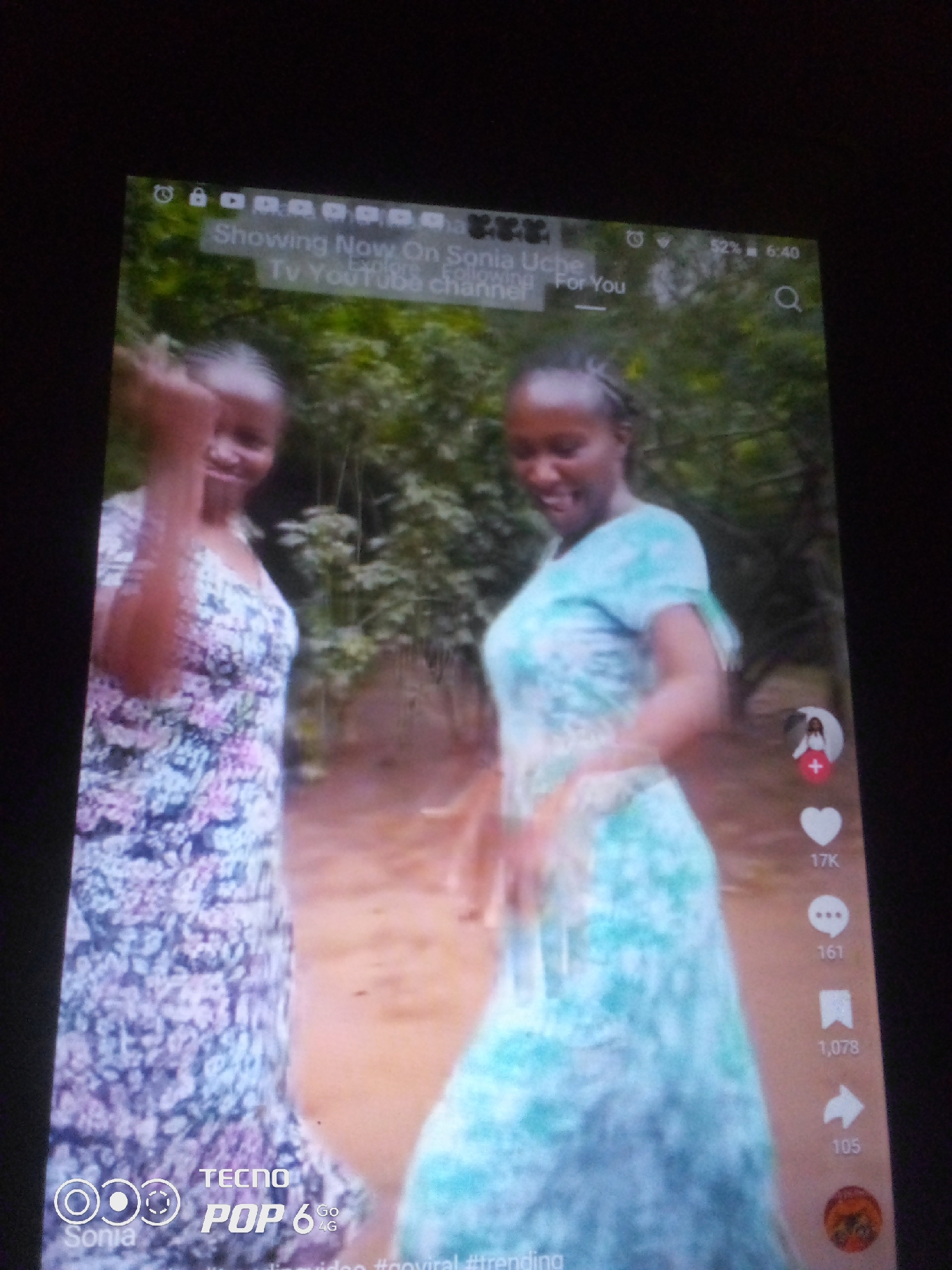 The image shows a close-up of a tablet screen displaying a video. Two young Black women, appearing to be in their late teens or early twenties, are visible. The woman on the left wears a floral print dress, and the woman on the right wears a light blue tie-dye dress. They are outdoors, with lush green foliage in the background, suggesting a rural or natural setting. The tablet screen displays typical social media interface elements, including a profile picture with a "+" symbol, a heart icon with "17K" below it, a speech bubble icon with "161" below it, a bookmark icon with "1,078" below it, and an arrow icon with "105" below it. Text overlays on the screen read "Showing Now On Sonia Uche," "Explore," "Follow," and "For You," along with "Tv YouTube channel." At the bottom of the screen, "TECNO POP 6 4G" and "Sonia" are visible, indicating the device and possibly the creator of the video. There is no indication of the specific city of Howayi, Nigeria, within the image itself.