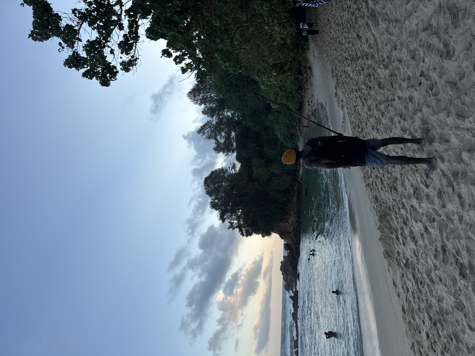 A man wearing a yellow beanie, a black tank top, and blue shorts stands on a sandy beach, facing the ocean. He is holding a long pole. The ocean is calm with several people swimming and wading. Rocky outcrops are visible in the water. Dense green foliage covers a hillside in the background. The sky is a soft blue with scattered clouds. The scene appears to be Goviyapana, Sri Lanka, during twilight.