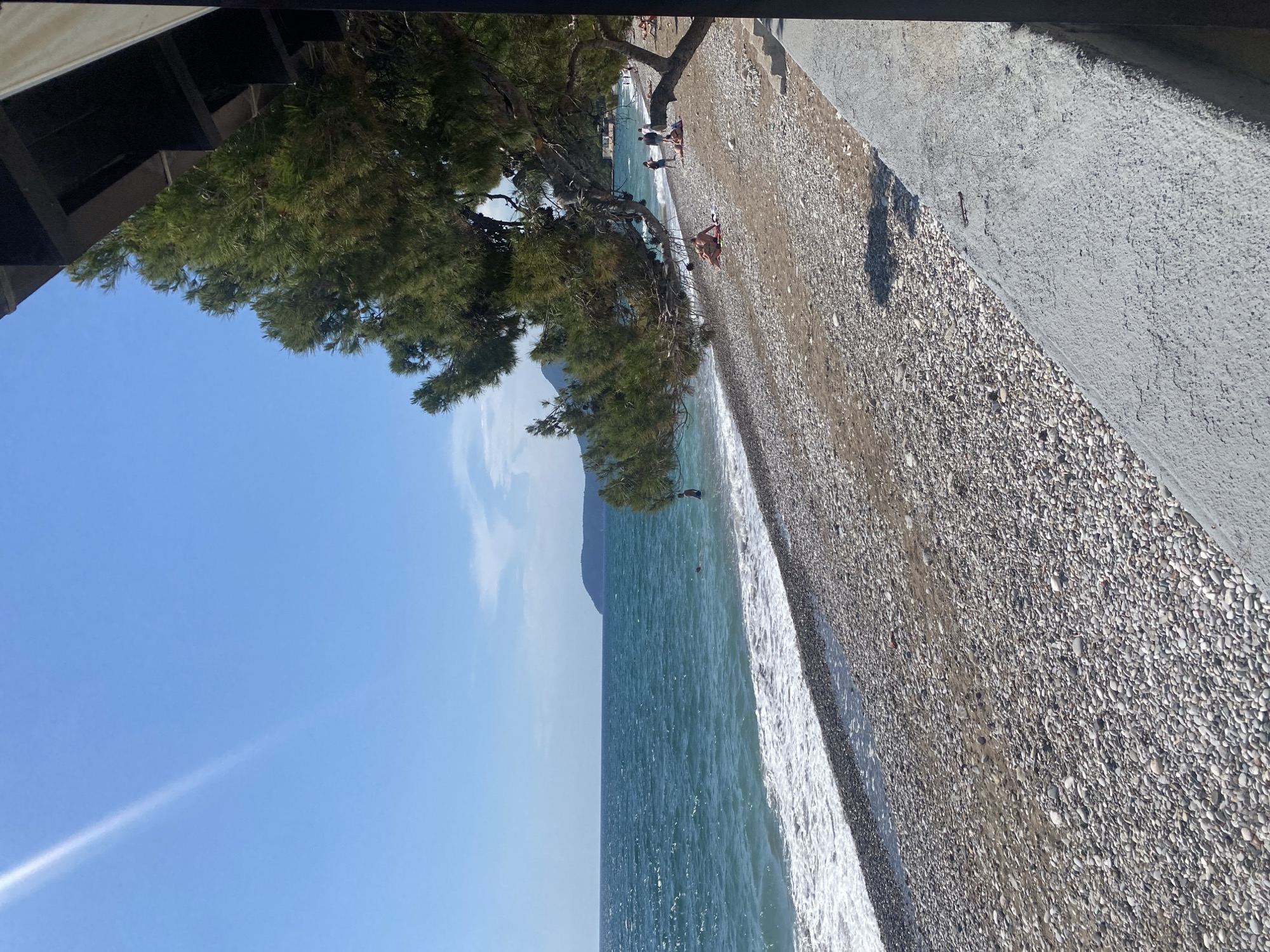 The image shows a beach scene in Bar, Montenegro, featuring a pebbled shoreline meeting the Adriatic Sea under a partly cloudy, blue sky. A large tree provides shade along the beach where several people are sunbathing and walking. A concrete structure borders the edge of the beach.