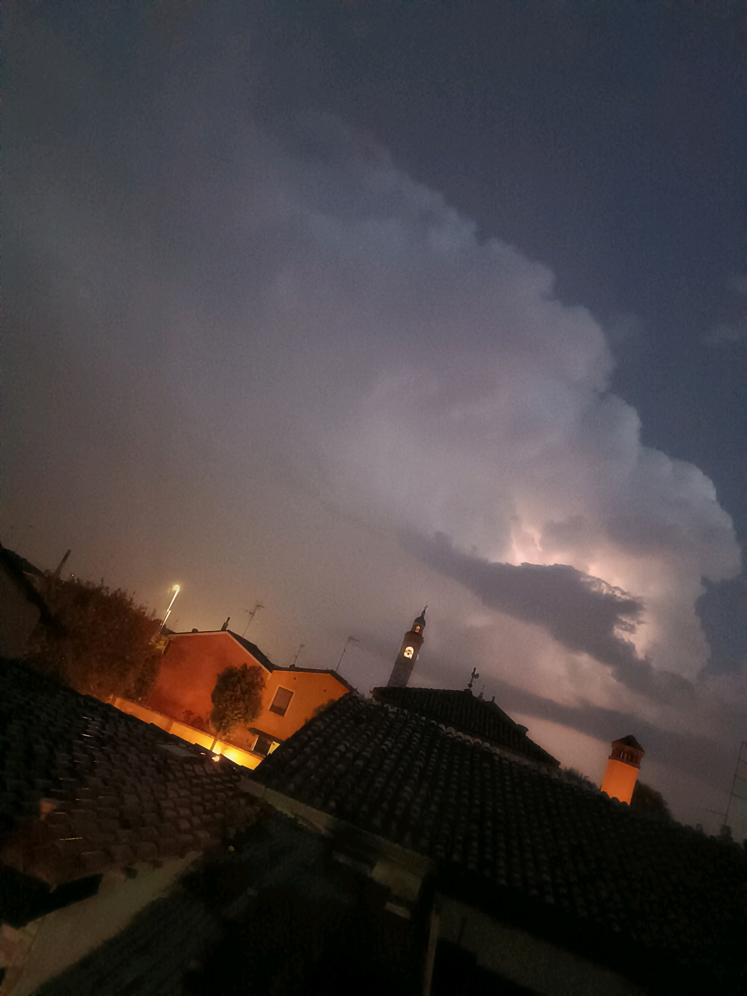 This image captures a dramatic thunderstorm over the city of Viadana, Italy. The sky is dark and ominous, filled with heavy clouds illuminated by a bright lightning strike in the distance. In the foreground, the tiled rooftops of houses are visible, casting shadows. Among the buildings, a bell tower with a lit clock face stands out, and another building features a chimney that is brightly lit from within, suggesting an interior light source. The presence of streetlights and antennas also indicates an urban environment. The overall atmosphere is one of a powerful natural event occurring within a populated area.