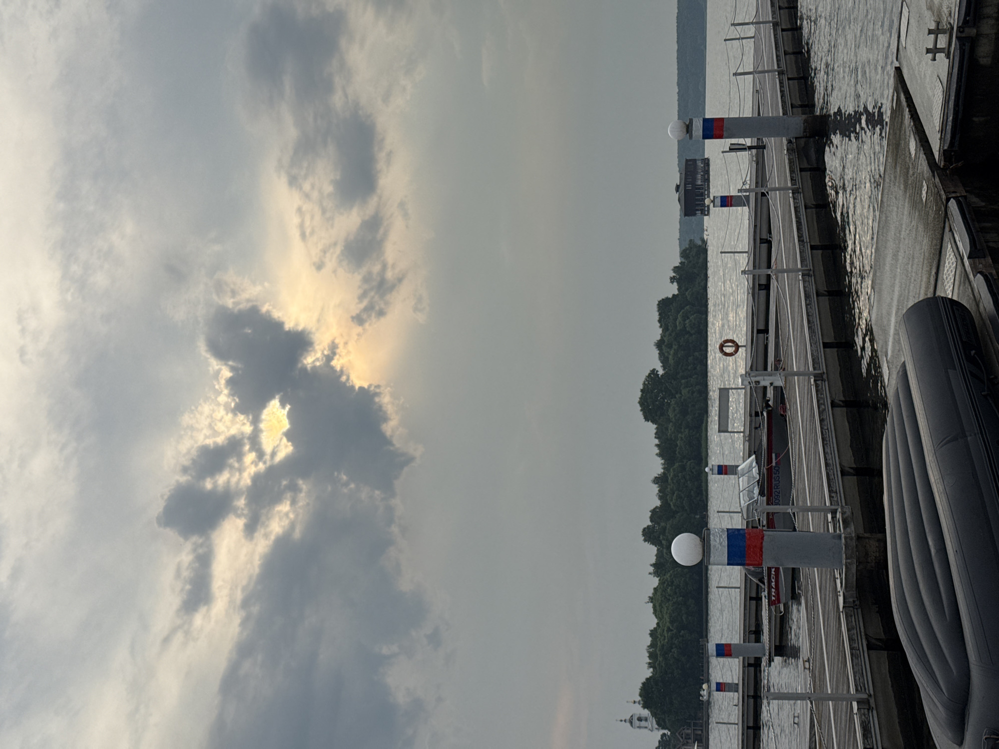 The image depicts a view of a wooden pier extending into a body of water under a cloudy sky. The sun is partially visible through the clouds, casting a soft glow. Several boats are docked at the pier, and there are cylindrical posts with red, white, and blue stripes, consistent with Russian flag colors, marking the pier's edge. In the distance, a treed island with a building and a church spire is visible on the left, and another building on stilts stands in the water on the right. The foreground features an inflatable boat lying on the pier. The scene appears to be set in a peaceful, possibly recreational area on the outskirts of the city of Троицкое, Russia, likely a lakeside or riverfront location.