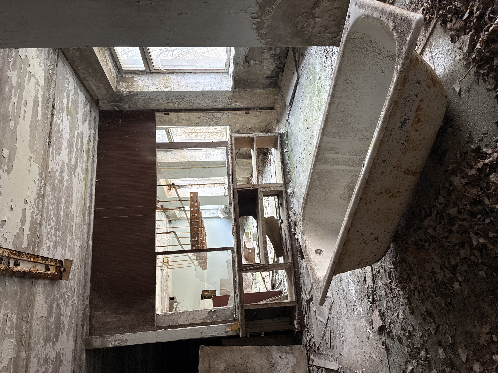An interior shot shows a derelict room in a building in Prypiat, Ukraine. The room is decaying and littered with debris. A white bathtub is positioned at an odd angle against one wall. An open, dilapidated frame, perhaps for a shelf or cabinet, sits askew in the center of the shot. Beyond that is another room visible through a doorway, and metal shelving units are seen in that next room. The walls are heavily damaged with peeling paint. There is a skylight in the roof, illuminating the scene. The floor is covered in a layer of leaves and debris. A rusted metal bar is affixed to the left wall.