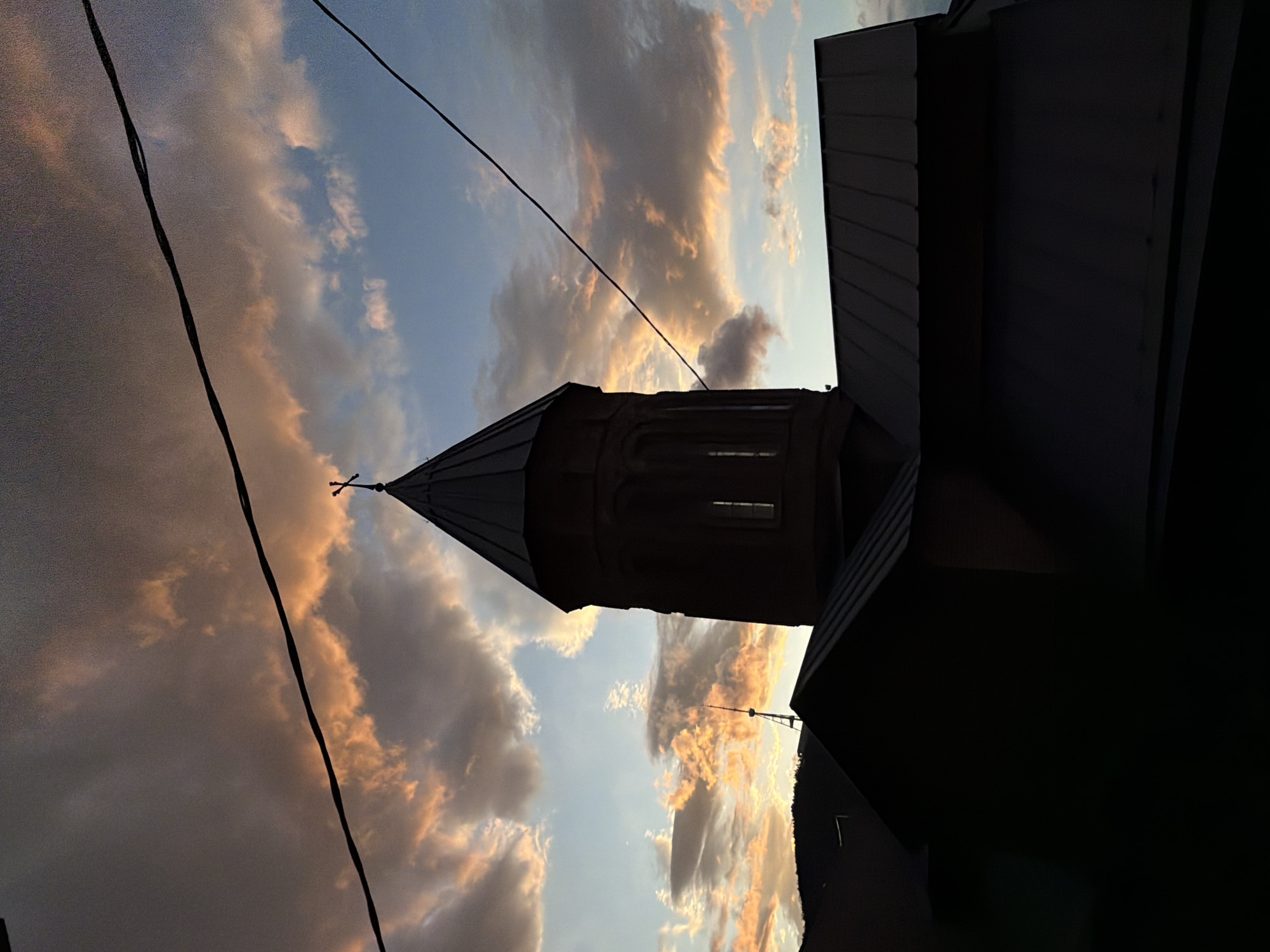 The image, rotated 90 degrees, depicts the Anchiskhati Basilica in Tbilisi, Georgia, against a cloudy sky at sunset. The basilica's tower, topped with a cross, is silhouetted against the sky. Power lines run diagonally across the frame. The surrounding buildings are dark and mostly obscured. The sky shows a gradient of blue and orange hues, indicative of sunset light reflecting off the clouds. The scene is static and shows no observable actions or interactions.