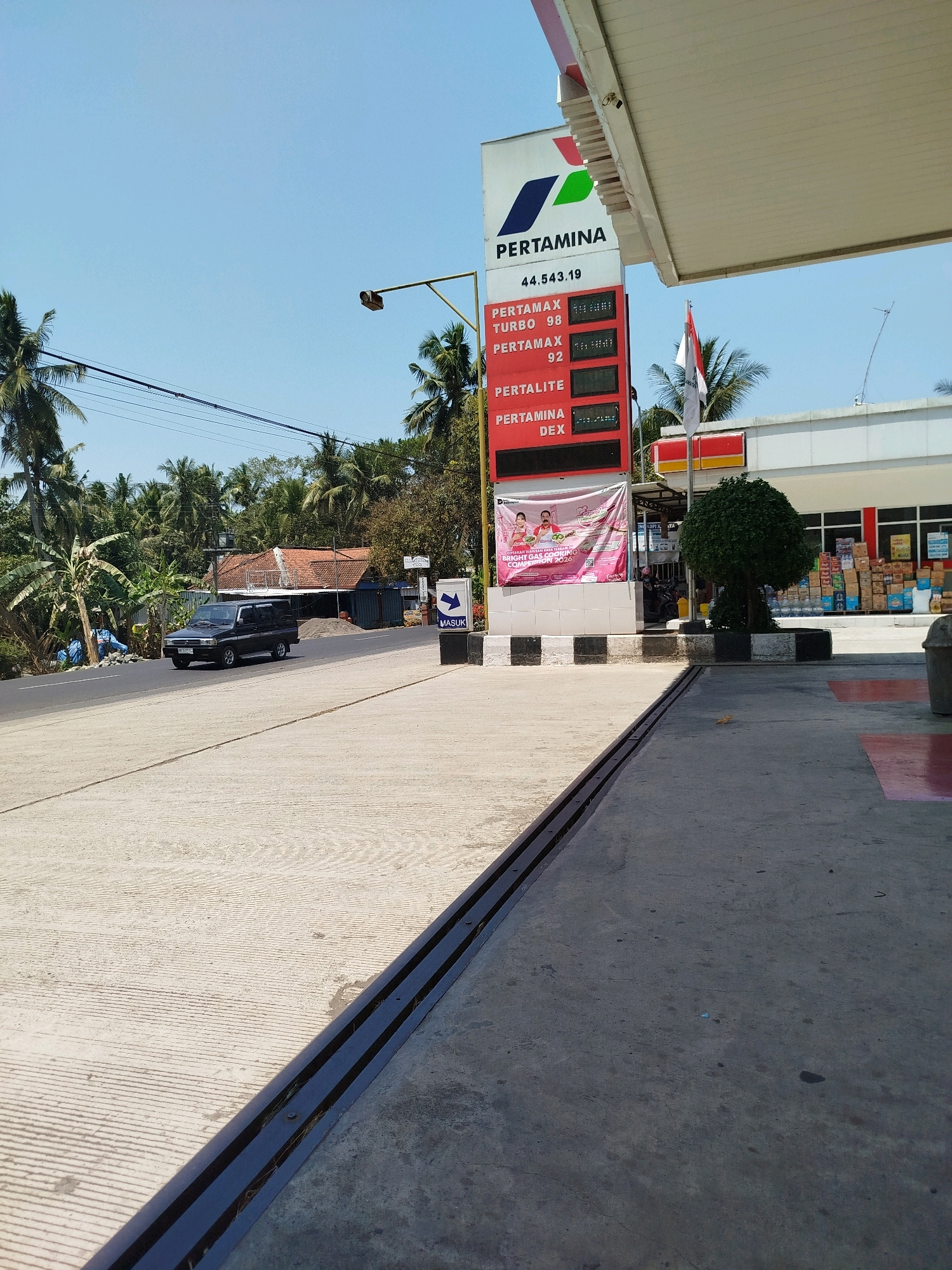 The image depicts a gas station in Bocor, Indonesia, on a sunny day. A Pertamina gas station sign is prominently displayed, showing fuel prices for various types of gasoline, including Pertamax Turbo, Pertamax, Pertalite, and Pertamina Dex. The prices displayed are: Pertamax Turbo 98 at 14,000, Pertamax 92 at 13,500, Pertalite at 10,450, and Pertamina Dex at 13,850. The total for Pertamax is listed as 44,543.19. A black car is driving on the road in front of the gas station. A banner promoting a "Bright Gas Cooking Competition 2016" is displayed on the side of the gas station. The environment includes palm trees, a building that appears to be a convenience store, and a street. The foreground shows a concrete surface with a metal drainage grate.