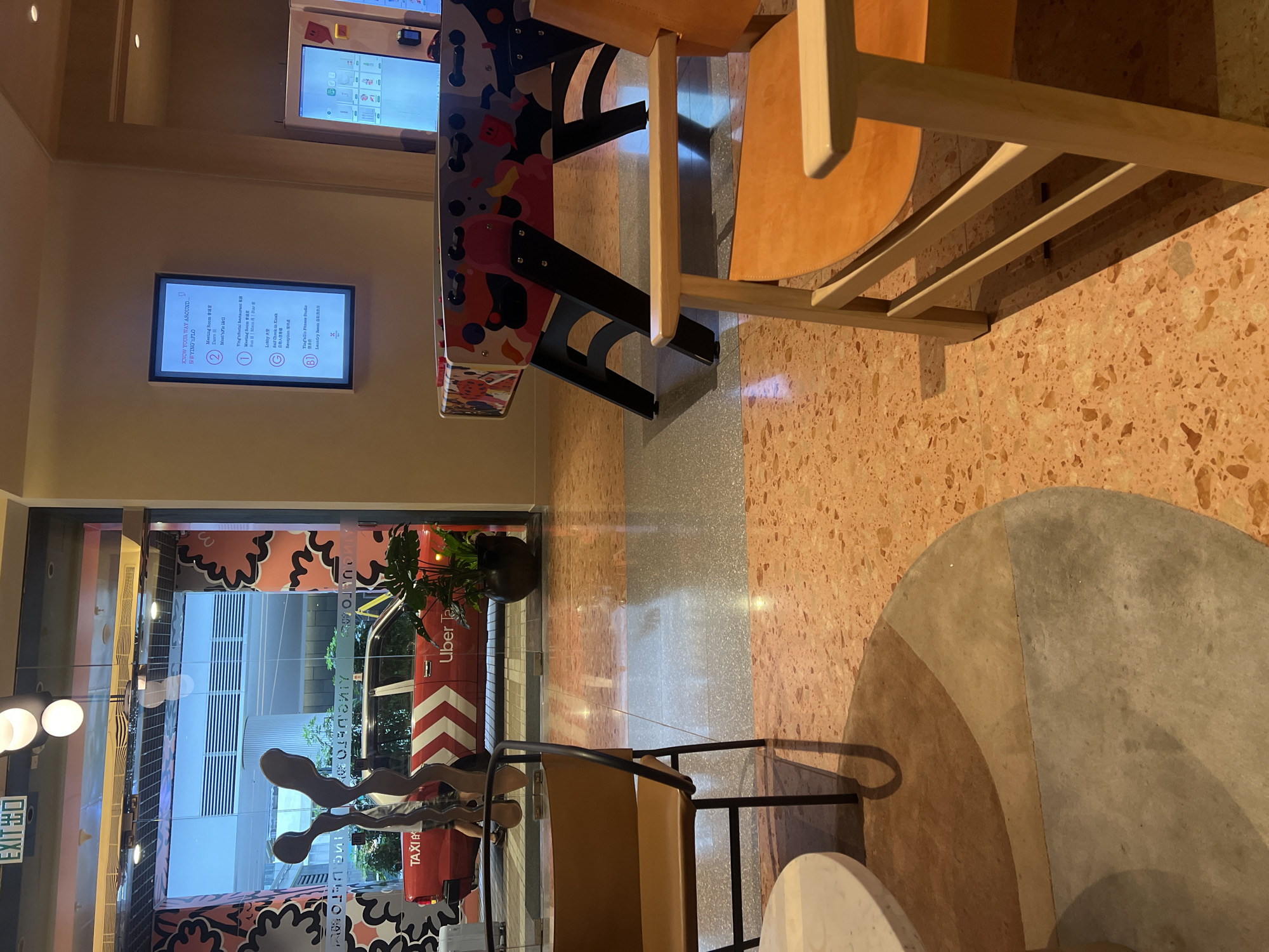 The image depicts the lobby of a hotel in Hong Kong, China. In the foreground, an orange leather armchair with a light-colored wooden frame is positioned on a terrazzo floor with an orange and grey pattern. To the left, a portion of a round marble-topped table is visible. In the mid-ground, a foosball table with a colorful, abstract design rests on the terrazzo floor. Beyond the foosball table, through a glass doorway, a red Uber taxi is parked outside. The taxi has "TAXI" and "Uber Taxi" branding visible. A person is partially visible walking towards the taxi. A potted plant with large green leaves is situated to the right of the taxi. In the background, the exterior of a modern building with horizontal window blinds is visible. The overall scene suggests a contemporary and stylish hotel interior with a playful element. The lighting suggests it is daytime.