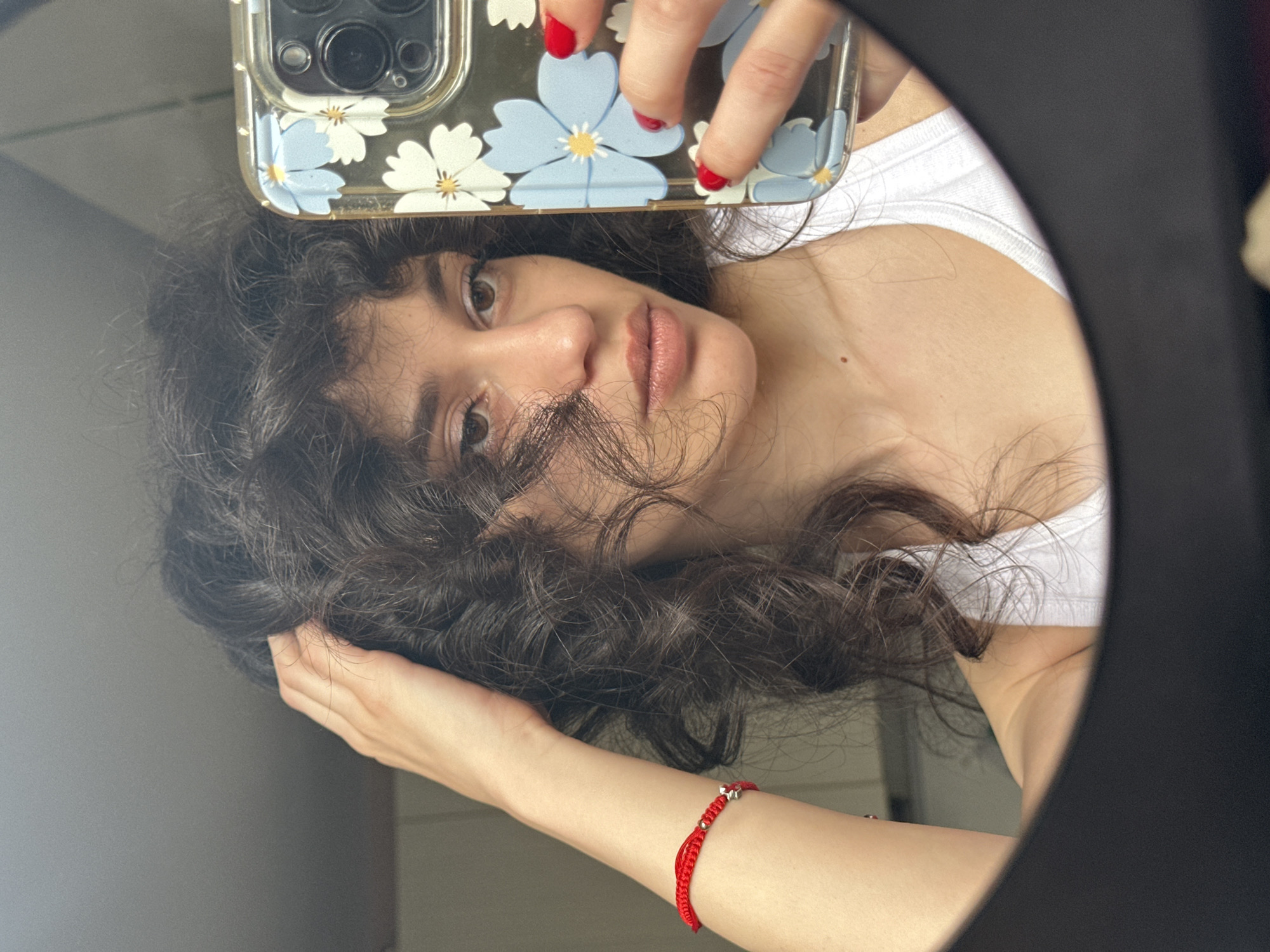 A woman is reflected in a mirror, holding her curly, dark hair with her left hand. She is wearing a white tank top and a red string bracelet on her left wrist. Her right hand holds a phone with a floral case, which is also reflected in the mirror. The background appears to be an indoor setting, possibly a room in a building. The image was taken in Sofia, Bulgaria.