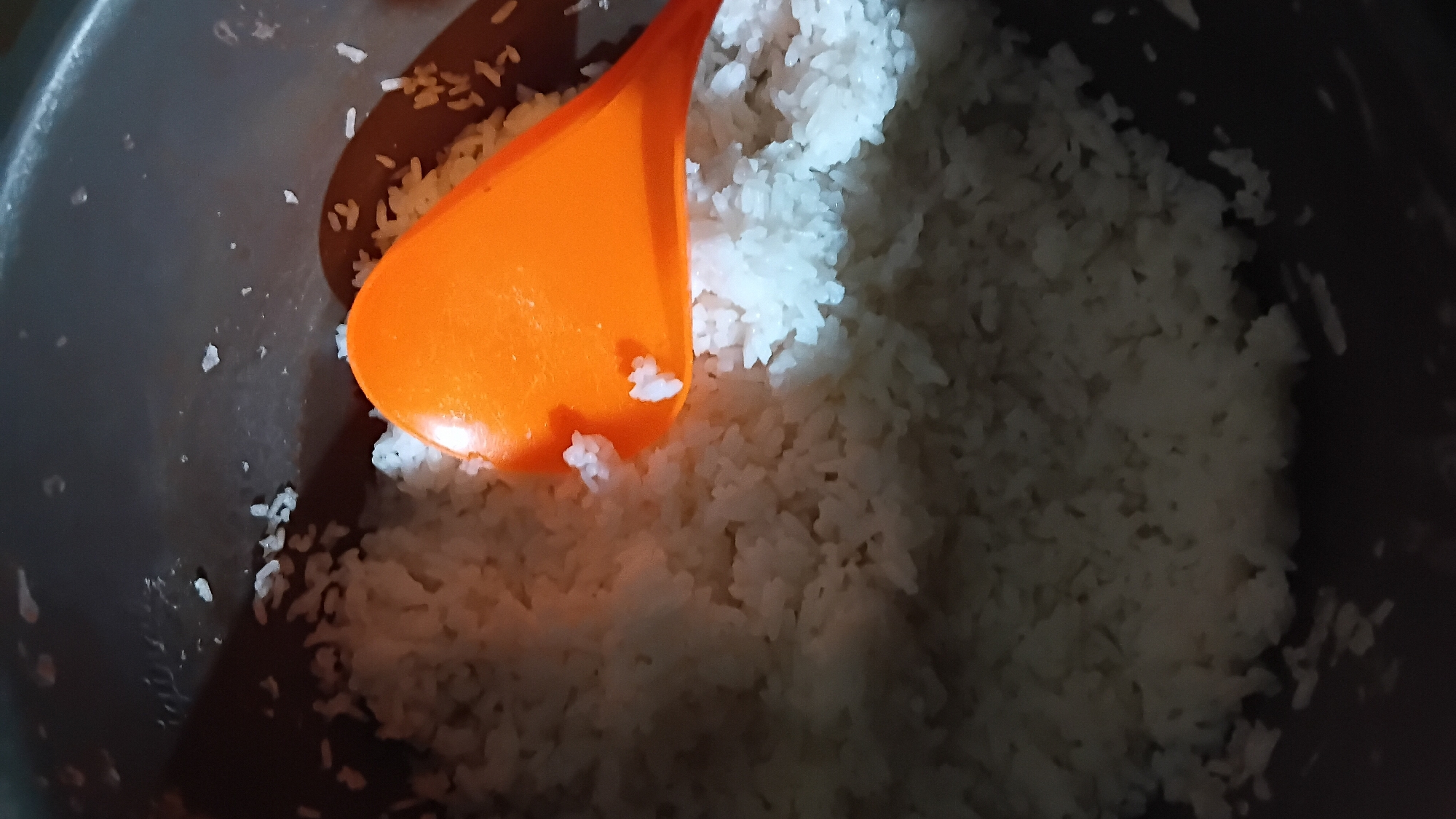 A close-up, high-angle shot shows cooked white rice in a dark pot, with an orange plastic rice paddle resting on top of the rice. The rice fills most of the pot, with individual grains visible. Small specks of rice are scattered around the rim of the pot. The scene appears to be a kitchen setting in Prabumulih, Indonesia.