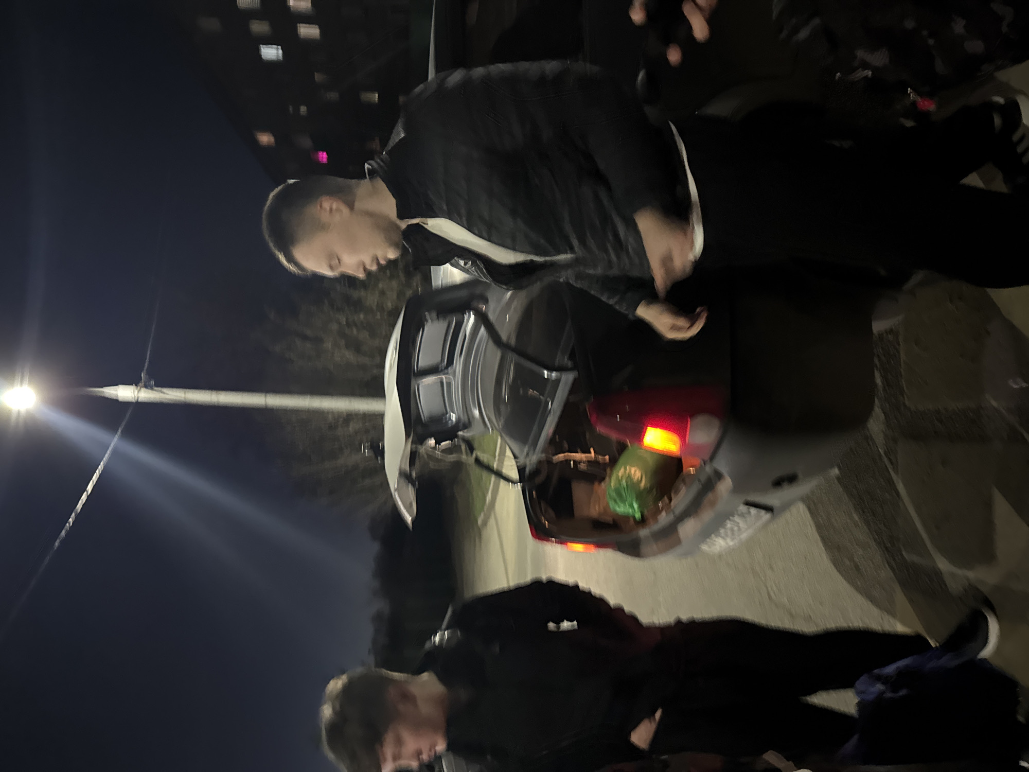 This image depicts an outdoor night scene, likely in Kazan, Russia, centered around two men and a car. The primary subject in the mid-ground is a man with short, dark hair, wearing a dark jacket with white accents. He is seen from the waist up, leaning slightly forward and looking towards the open trunk of a dark-colored sedan. His hands are positioned as if he is interacting with items inside the trunk. Visible within the car's trunk is a bright green plastic bag. The car's red taillights are illuminated.
In the lower foreground, partially obscured, is another man with short, dark hair, dressed in dark attire. He appears to be standing near the car, looking downwards.
The setting is dark, characteristic of nighttime, with artificial light sources providing illumination. A prominent streetlight on the left side of the frame emits strong rays, creating a visible lens flare. In the background, a building is visible with several illuminated windows, casting rectangular glows of pink and yellow light. The ground beneath the subjects and the car is a dark, paved surface.
The activity taking place appears to be the loading or unloading of items from the car's trunk. The overall mood is neutral, with no strong emotions discernible from the individuals' expressions. No visible text can be identified in the image.