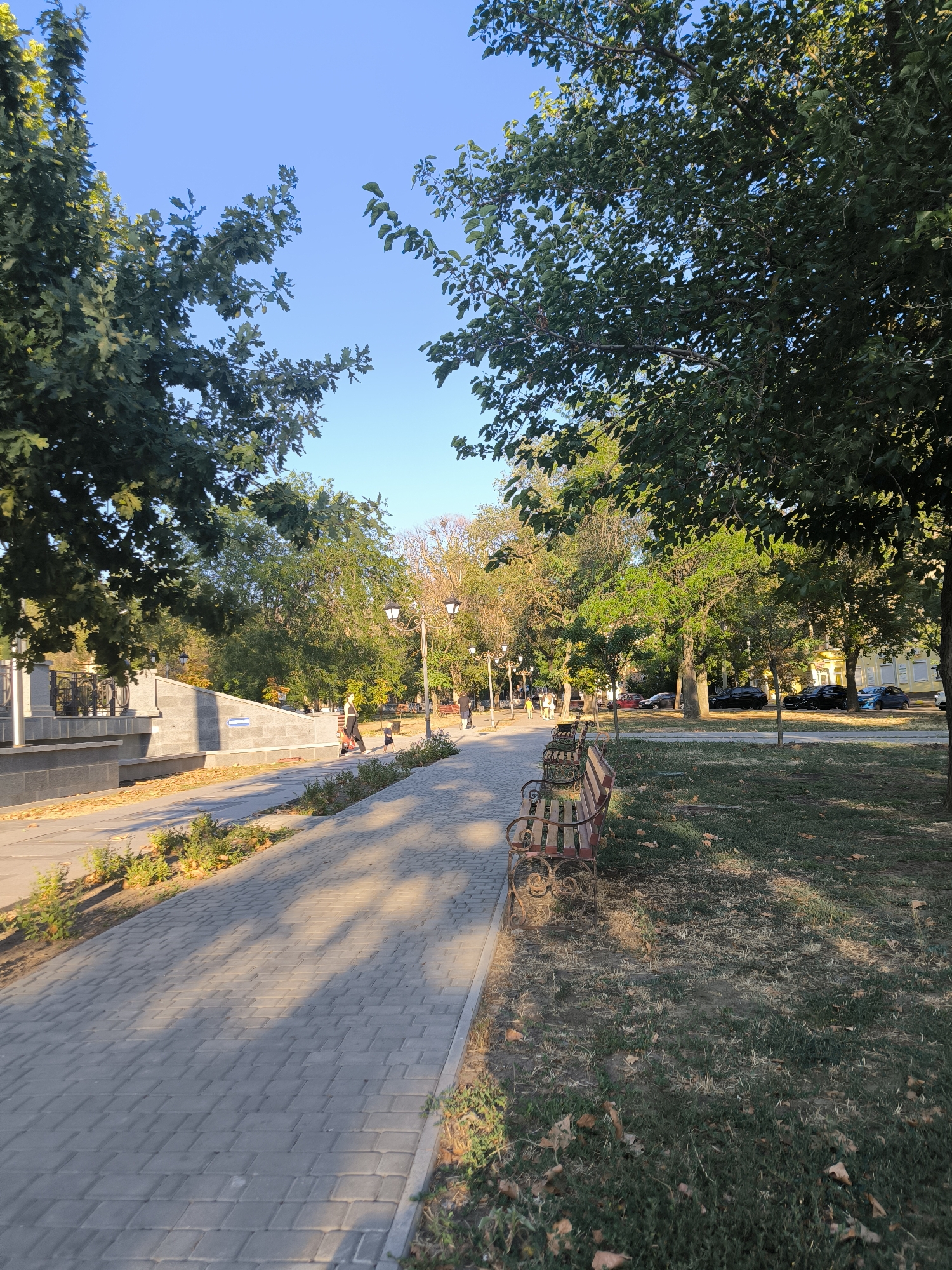 The image depicts a sun-dappled park pathway in Mykolaiv, Ukraine. The pathway, paved with gray rectangular bricks, runs diagonally from the lower left to the upper right. To the left of the pathway is a raised, gray stone structure with decorative metal railings. On the pathway, a person is walking with a child. To the right of the pathway, a grassy area contains several park benches with decorative metal frames and wooden slats, some of which are empty. Trees with green leaves line the pathway and the grassy area, with their branches extending into the clear blue sky. In the background, streetlights are visible, and further back, buildings and parked cars can be seen. The overall atmosphere is peaceful and idyllic, suggesting a late afternoon in the park.