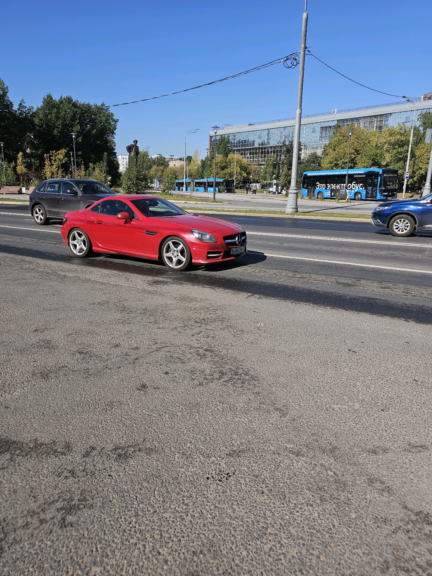 A red Mercedes-Benz convertible is parked on the side of a city street in Moscow, Russia, possibly on a service road or shoulder. The car is facing left, with its front end angled slightly towards the camera. The license plate is partially obscured but appears to have Cyrillic characters. Behind the Mercedes, a dark-colored SUV is also parked. Further down the road, a blue electric bus is visible with the text "Это электробус" (This is an electric bus) written on its side. The background features a modern building with many windows, trees with autumn foliage, and a clear blue sky. The road surface appears to be asphalt with some patches and stains. A lamppost with wires is in the foreground. There are no people clearly visible interacting with the vehicles.
