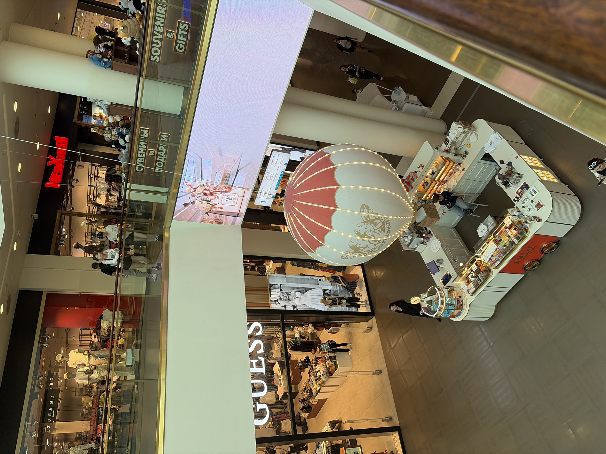 An overhead, medium shot captures a bright, multi-level shopping mall interior. In the foreground, a large, decorative hot air balloon, illuminated by tiny white lights, hangs suspended from the ceiling. Below the balloon, a small, white kiosk with red accents sells sweets. It features a glass display case filled with colorful candies and chocolates, and a rotating carousel display of smaller items on top. A young woman with dark hair, wearing a black jacket, stands to the left of the kiosk, looking towards it. Another person is seated on a stool behind the counter, and others are visible further into the mall.
On a mid-level balcony to the left, the exterior of a "GUESS" clothing store is visible, with mannequins dressed in casual wear displayed in the windows. Shoppers can be seen browsing inside. Further back, other storefronts are present, including one with a prominent red "NEW YORKER" logo. Signs in both Russian and English, indicating "SOUVENIRS & GIFTS," are visible on the railings of the balconies. The mall appears to be modern and well-lit, with polished floors and extensive use of glass and metal in its architecture. The overall impression is one of a busy, commercial environment. The presence of Russian signage and the architectural style suggests the location is likely a shopping mall in Saint Petersburg, Russia.