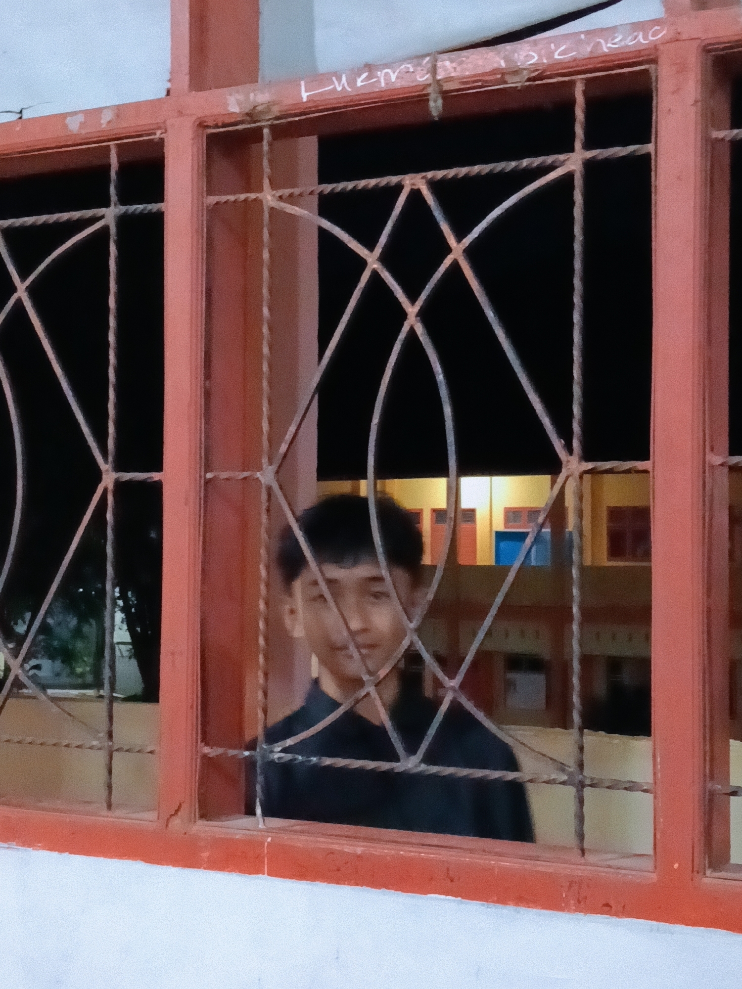 A person stands behind a red-orange window grill. The person has short dark hair, appears to be young, and is wearing a dark shirt. The window grill is composed of squares and curved diagonal wires. Outside the window, there is a building with multiple windows and red and blue colored elements, visible due to the night time. The building appears to be located in Lhokseumawe, Indonesia based on visual context.