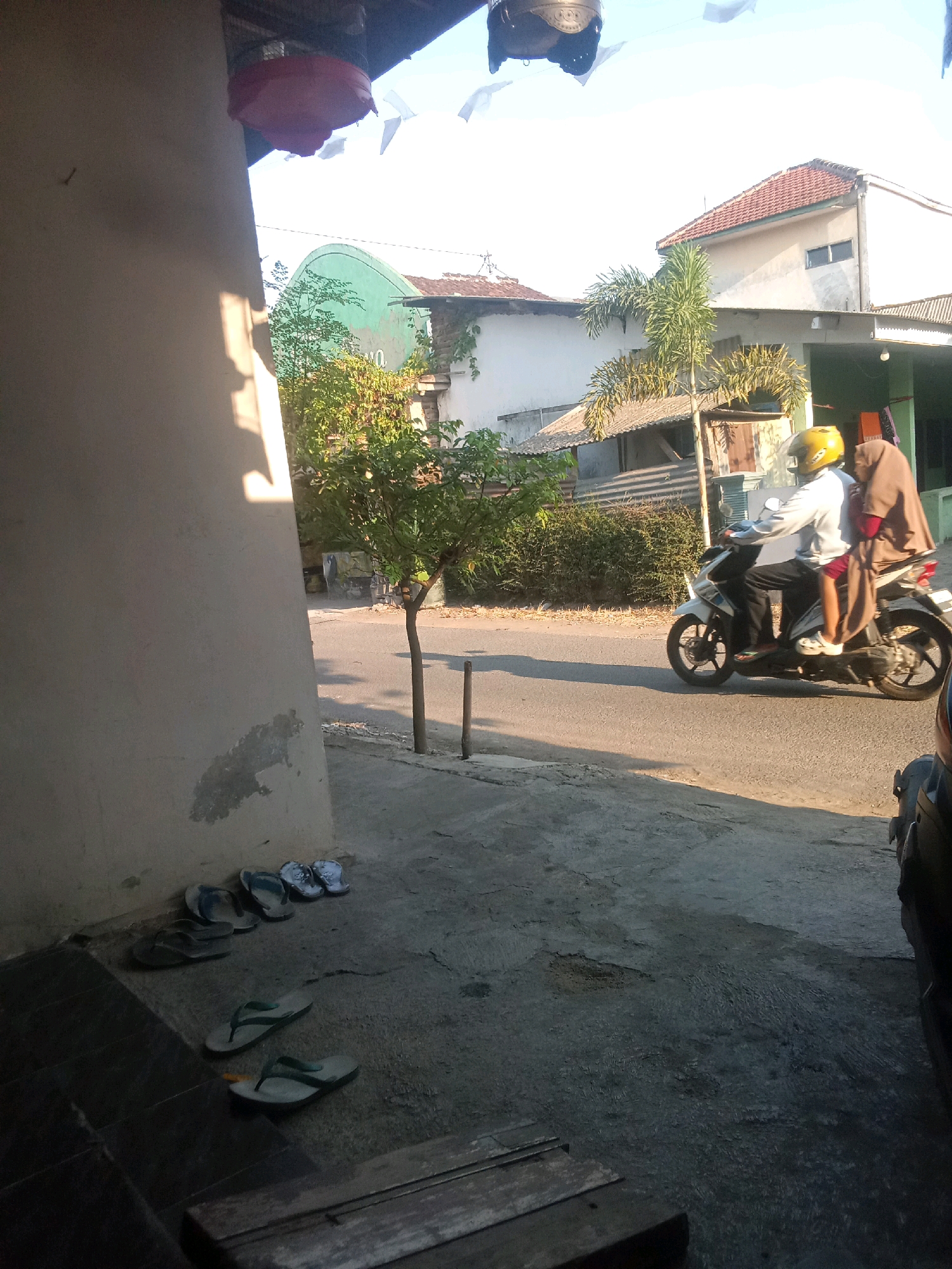 The image depicts a street scene in Jubung, Indonesia, bathed in the warm light of what appears to be early morning or late afternoon. In the foreground, several pairs of flip-flops are scattered on a textured concrete surface, suggesting a residential or casually maintained outdoor area. To the left, a light-colored wall with some patches of damage or discoloration dominates the frame, casting a shadow that stretches across the ground.
Further into the scene, a paved road runs horizontally. On this road, a white motorcycle is moving from left to right. The motorcycle carries two individuals. The driver, wearing a light-colored shirt and black pants, is wearing a yellow helmet. The passenger, a female wearing a brown headscarf and a maroon top, is seated behind the driver.
Behind the road, a small tree with green foliage stands prominently, its thin trunk supported by a thin vertical pole. Beyond the tree, houses with red-tiled roofs and light-colored walls are visible, indicative of Indonesian architecture. Vegetation, including bushes and possibly palm trees, frames the background. The sky is bright and clear, with hints of white decorative elements suspended from above in the upper left portion of the image. A dark object, possibly another vehicle, is partially visible on the right edge of the frame. The overall atmosphere suggests a typical day in a semi-urban or rural Indonesian setting.