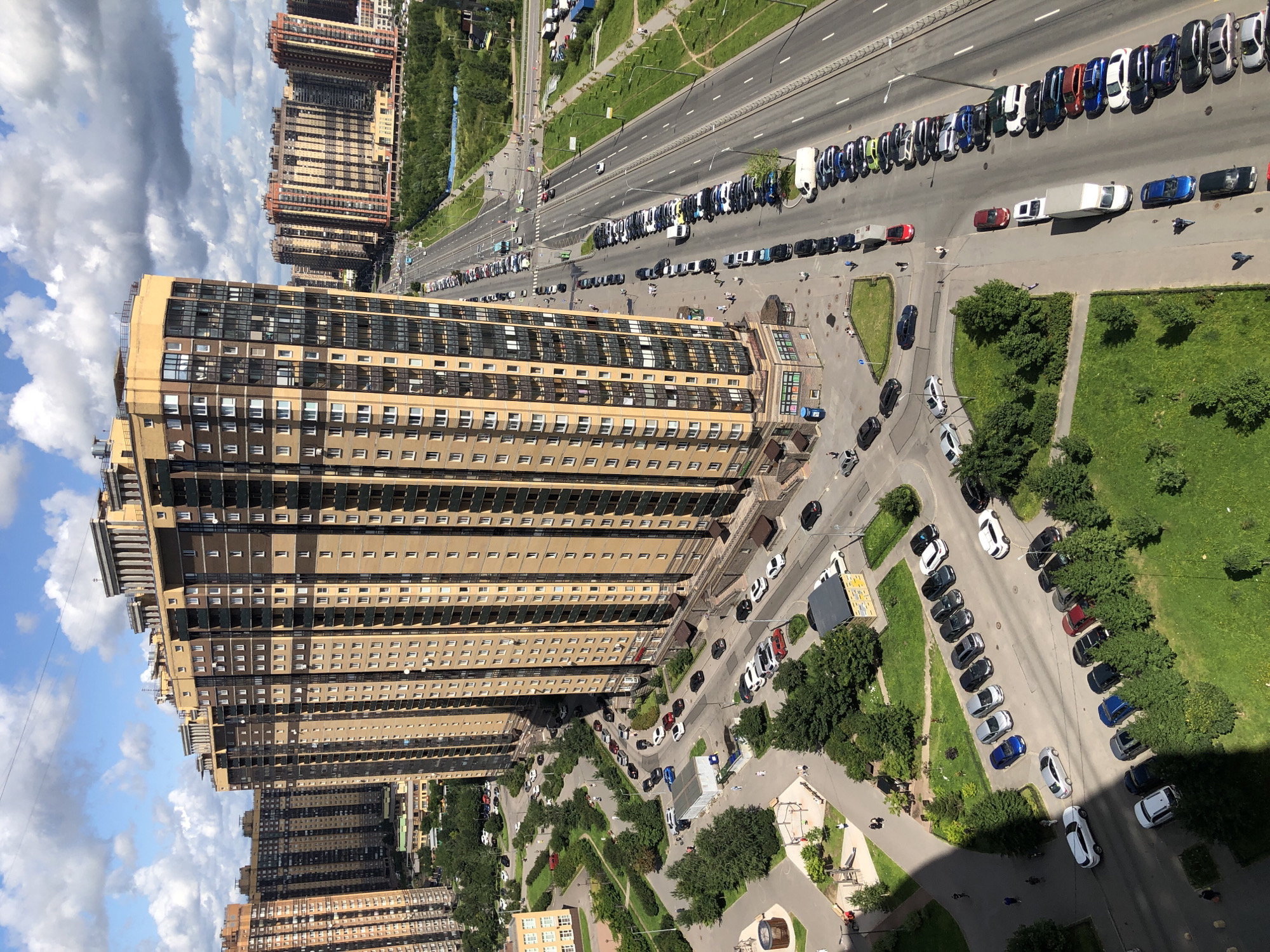 An aerial view captures a dense urban landscape dominated by towering residential buildings and a multi-lane road. The buildings, a mix of beige and brown tones, feature numerous windows and balconies, indicating high occupancy. The road, stretching across the frame, is lined with parked cars, suggesting a busy residential area. Green spaces, including trees and grass, are interspersed throughout the scene, offering a touch of nature amidst the concrete. The sky above is a mixture of blue and white clouds, hinting at fair weather. Several individuals are visible on the sidewalks and roads, engaged in various activities like walking and driving.