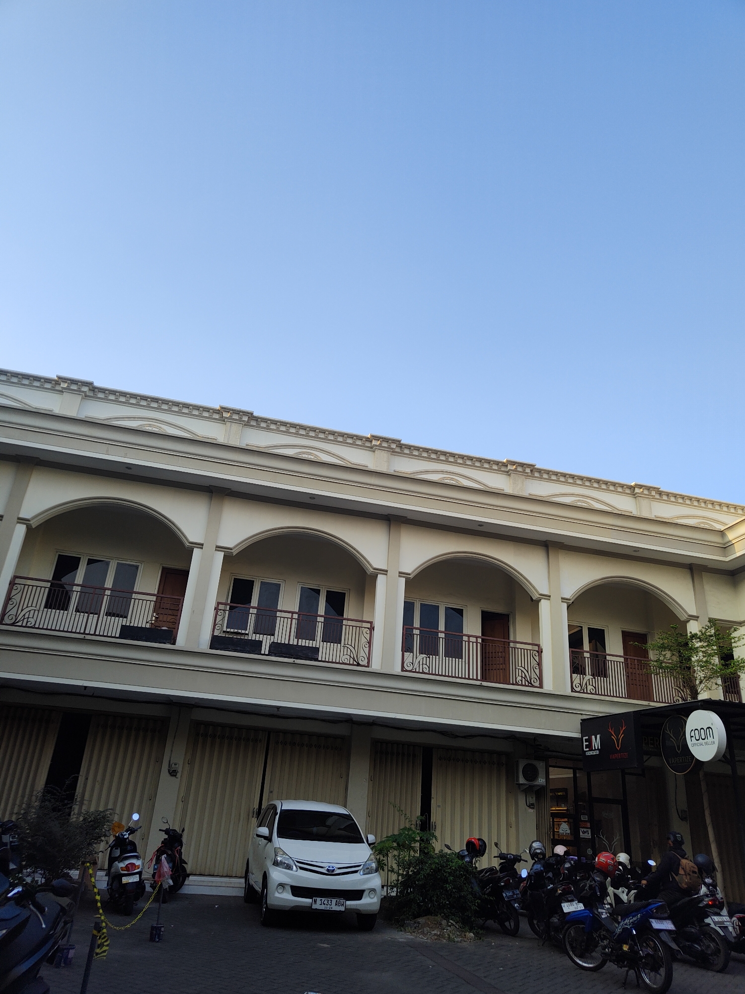 The image captures a view of a two-story building in Bangil, Indonesia, under a clear, pale blue sky. The building has a pale cream exterior with decorative moldings along the roofline. The second story features arched balconies with dark railings, each with multiple windows. The ground floor has large, closed metal roll-up doors. In front of the building, there is a white car parked, and several motorcycles are lined up. To the right, there are two shop signs: one that reads "EM VAPERTIZE" with a deer logo, and another circular sign labeled "foom OFFICIAL SELLER". A small green tree is visible to the right of the building. The overall scene depicts a commercial or residential street in a town.