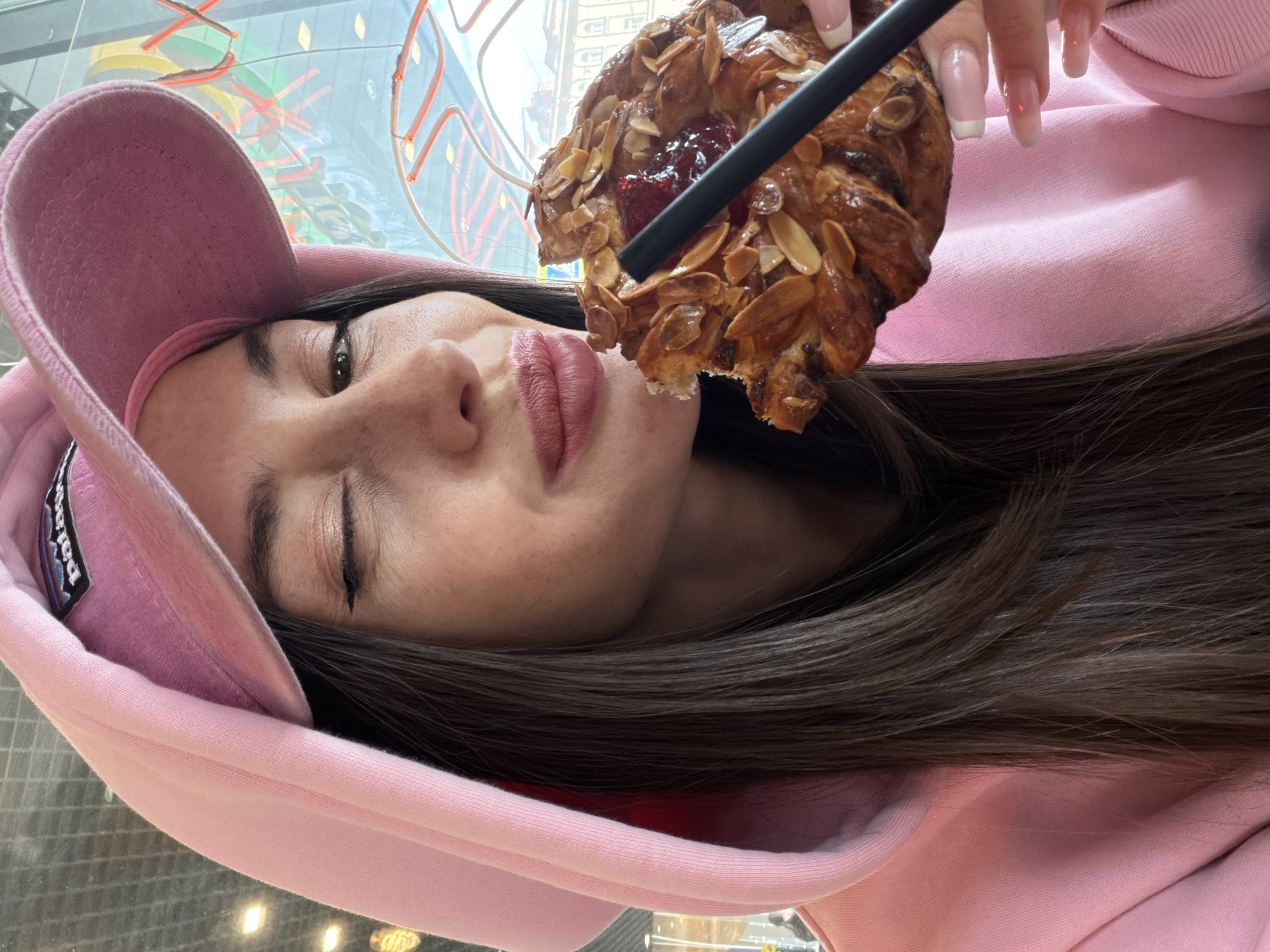 A young woman in a pink hoodie and cap holds a croissant with almond flakes and jam. The image is a selfie, with the woman positioned close to the camera. She is holding a black straw in the croissant. Her left eye is partially closed, and she has light pink lipstick. The background suggests an indoor setting near a window with colorful, abstract art possibly hanging outside. The scene is in Saint Petersburg, Russia, based on the style of architecture and the art visible through the window.