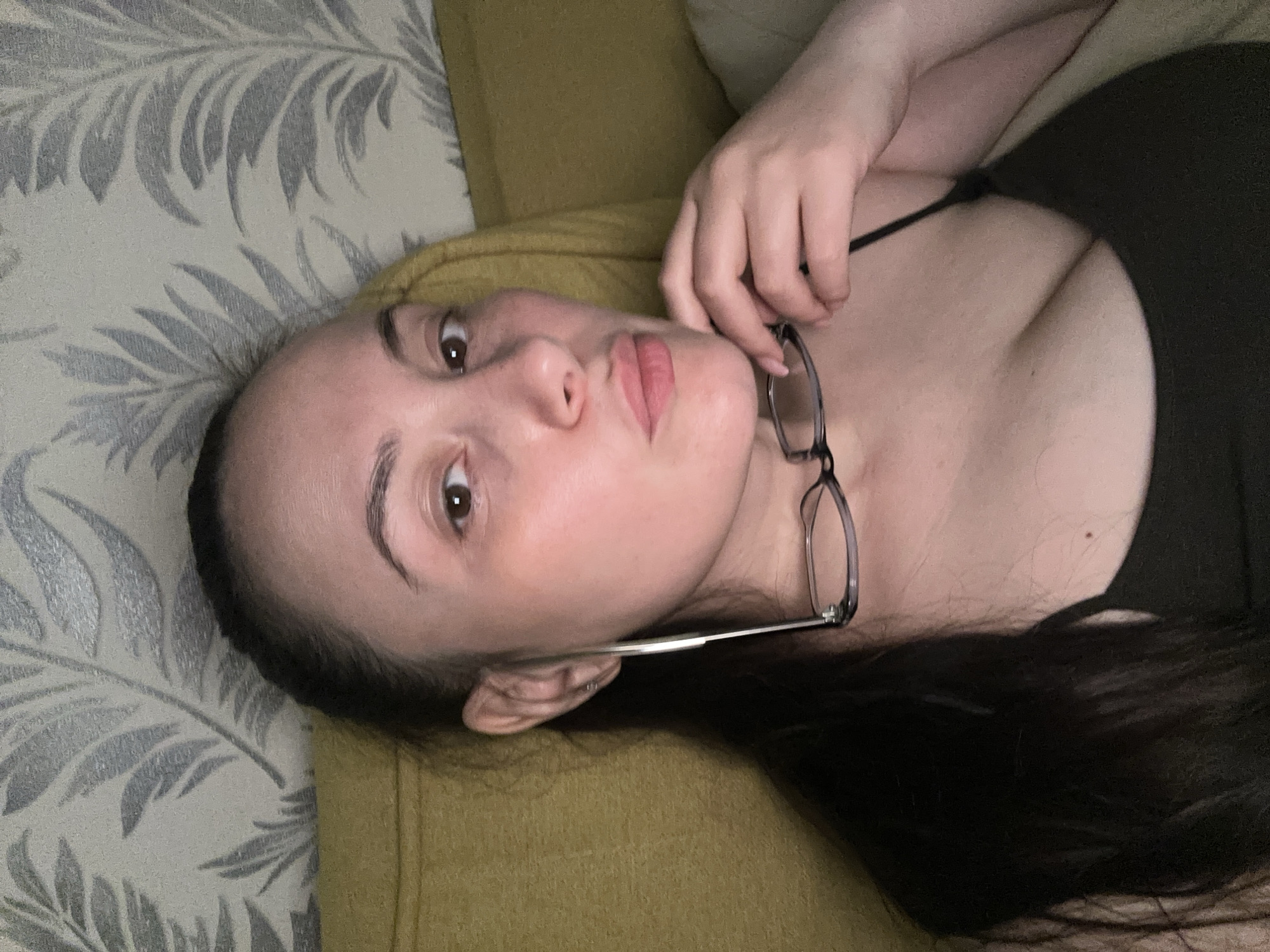 A woman is lying down on a surface covered in beige and grey patterned fabric. She is wearing a black top and holding a pair of glasses. She has long dark hair and is looking at the camera with a slight pout. The image was taken indoors. There is no information to determine if the image was taken in Chaikovsky, Russia.