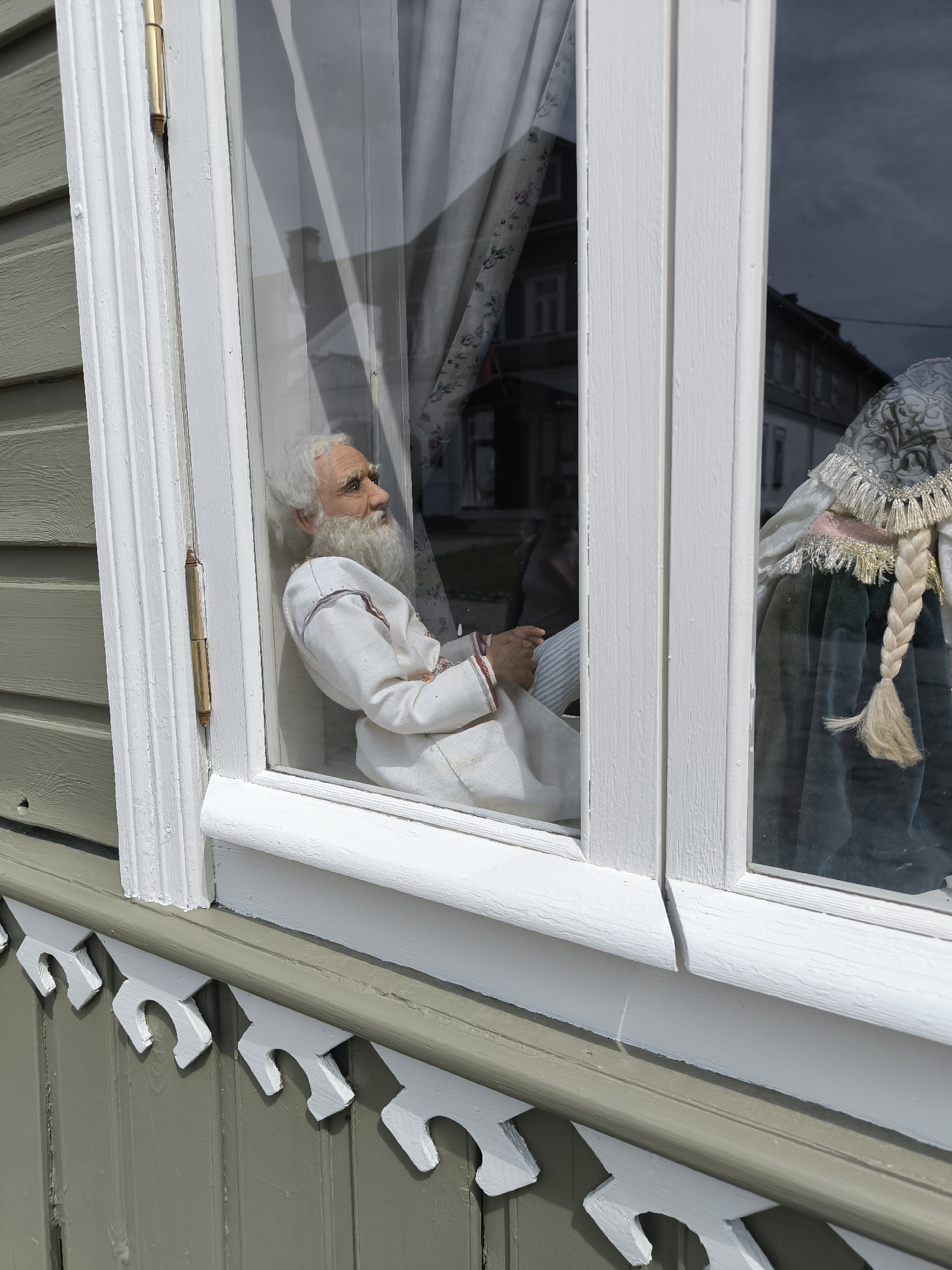 The image shows a close-up view of a window on the exterior of a building. The building has horizontal grey siding and decorative white trim along the bottom edge of the window. Inside the window, there are two mannequins. The mannequin on the left is an elderly man with white hair and a beard, wearing a white robe with red trim. He is seated and looking to the right. The mannequin on the right is a woman with a long blonde braid, wearing a dark green dress and a patterned headscarf with fringe. The window reflects the sky and buildings across the street, suggesting an outdoor setting in a town or city. The presence of traditional clothing on the mannequins may indicate a cultural or historical exhibit. Pechory, Russia is known for its historical sites and religious significance.