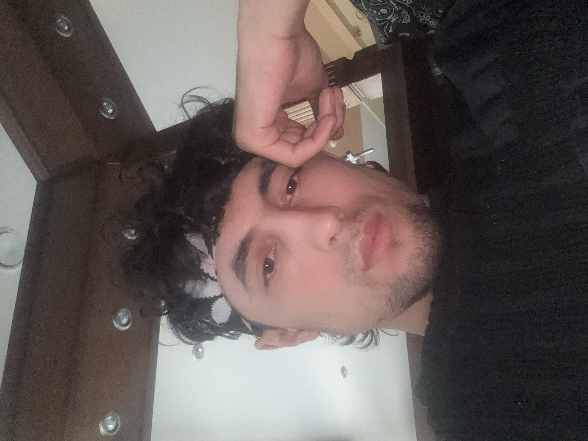 A close-up, sideways shot features a young person resting their forehead on their hand. The person has dark, curly hair, a bandana tied around their forehead, and is looking towards the camera. Their eyes are open and appear to be brown. They have a light beard and their lips are slightly parted. The background is blurred but appears to be indoors, with a wooden structure and a white wall visible. The lighting suggests it might be daytime, but the specific time of day is unclear. The overall mood seems relaxed or contemplative. No visible text is present in the image.
