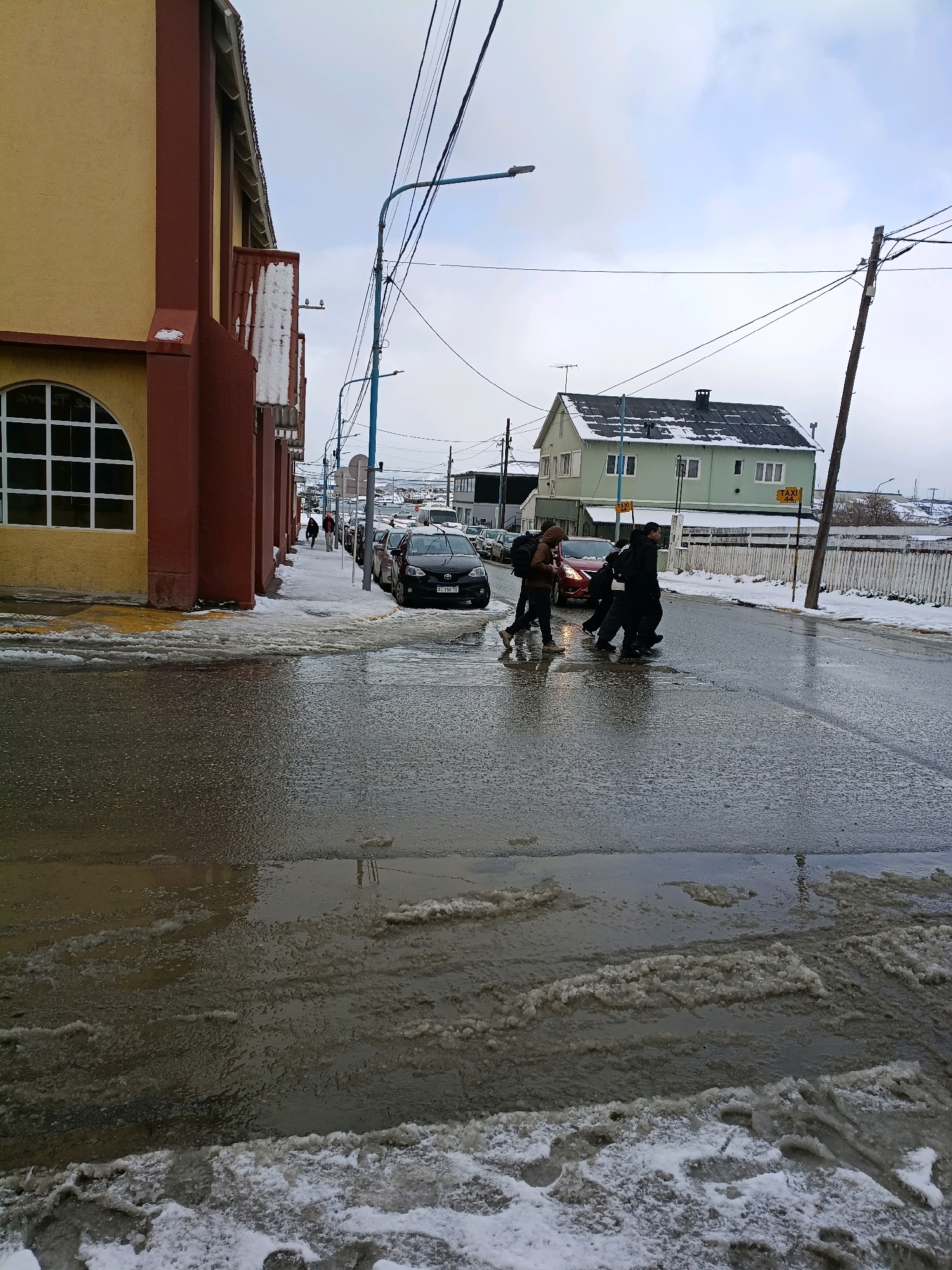 A wide-angle shot captures a city street in Ushuaia, Argentina, under an overcast sky, with patches of snow and melting ice on the road and sidewalk. In the foreground, the wet asphalt is covered in icy puddles and slushy snow. A building with a yellow facade and dark red trim lines the left side of the street, featuring a large arched window with white mullions. Across the street, a light green house with a dark roof stands out, with icicles hanging from the eaves. Several vehicles are parked along the right side of the street.
In the middle ground, three individuals are crossing the street. The person on the far left wears a hooded jacket and backpack. The person in the middle is wearing dark clothing. The person on the far right, also in dark clothing, appears to be walking a step behind the other two. A red car is stopped at the crosswalk, likely yielding to the pedestrians. A black car is visible further down the street.
The background shows more buildings, power lines crisscrossing the sky, and a general sense of a cold, possibly wintery, day in Ushuaia. The presence of snow and ice on the ground suggests a low temperature. The overall scene depicts a typical urban street environment in Ushuaia during a cold season.