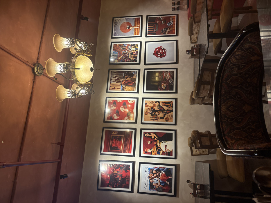 A wall displays twelve framed pictures in a grid arrangement, including images of a guitar, a dice, and the "Friends" television show cast. An ornate chandelier hangs from the ceiling nearby. A mirrored table reflects the chairs and interior decor. The setting is possibly a restaurant or lounge.