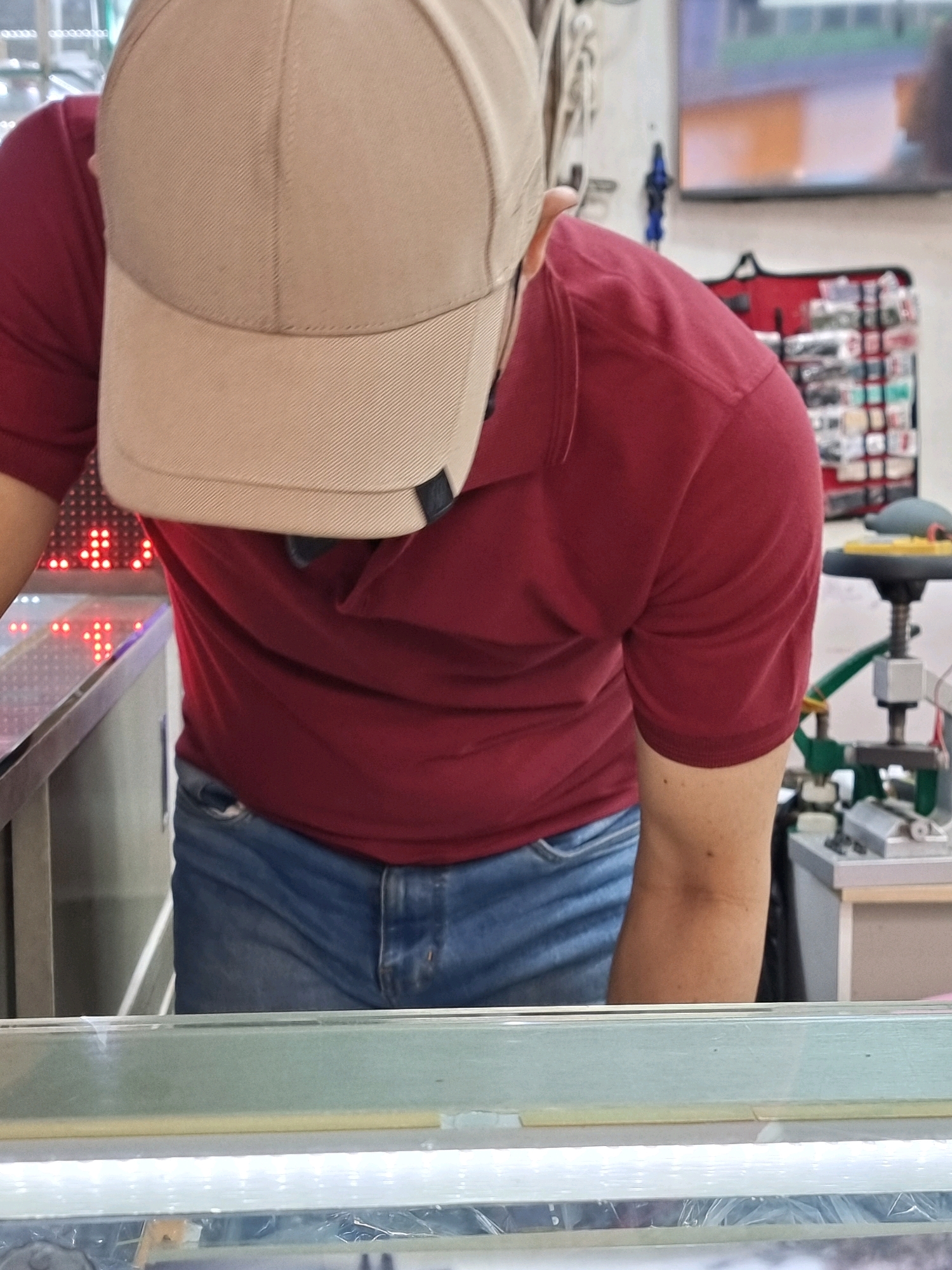 A person wearing a red t-shirt and blue jeans, and a beige baseball cap is standing in front of a counter with a display of red lights. In the background, a toolbox is visible. The scene appears to be indoors, possibly in a shop or workshop.