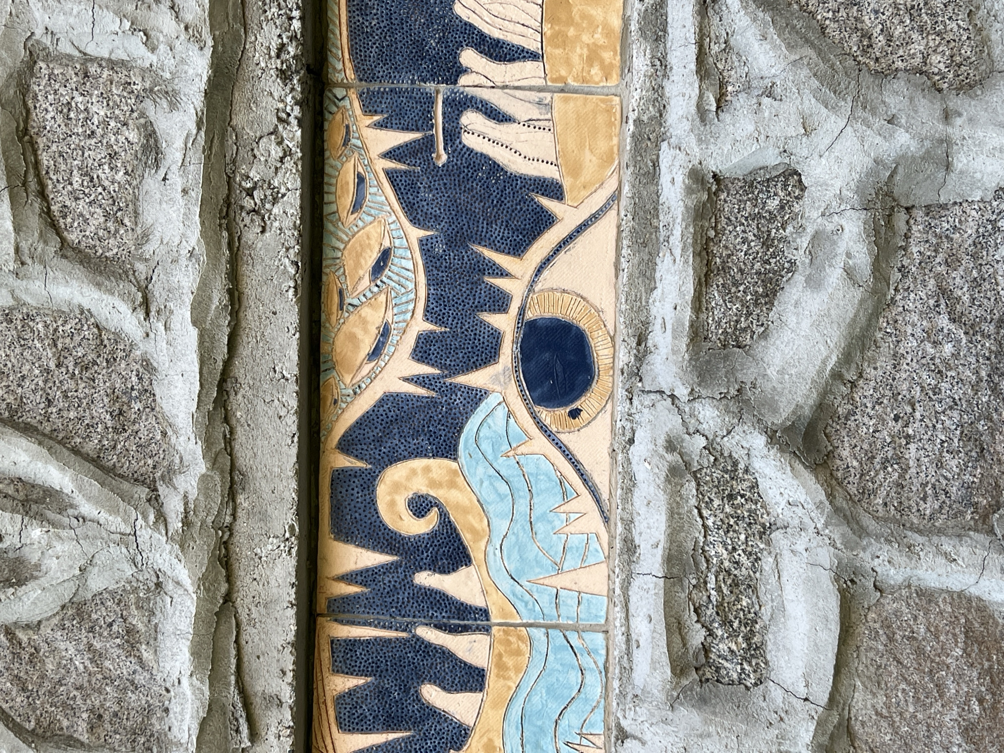 A close-up, eye-level, outdoor, medium shot shows a section of a textured stone wall with a decorative tile inlay. The wall is constructed of irregular, rough-hewn grey stones of various sizes, interspersed with cement mortar. The mortar is cracked and appears weathered. The tile inlay is a horizontal band, approximately 20cm high, featuring a stylized, abstract design with a large, prominent eye in the center. The eye has a dark blue pupil and is surrounded by pale yellow and blue wavy lines, possibly representing water or a dynamic element. Above the eye, the design continues with jagged yellow shapes and a dark blue background with fine speckles, reminiscent of teeth or a monstrous mouth. To the left of the eye, there are white, hand-like shapes emerging from the blue and yellow elements. To the right, there are more white, stylized shapes. The tile inlay appears to be part of the facade of a building located in Sinemorets, Bulgaria.