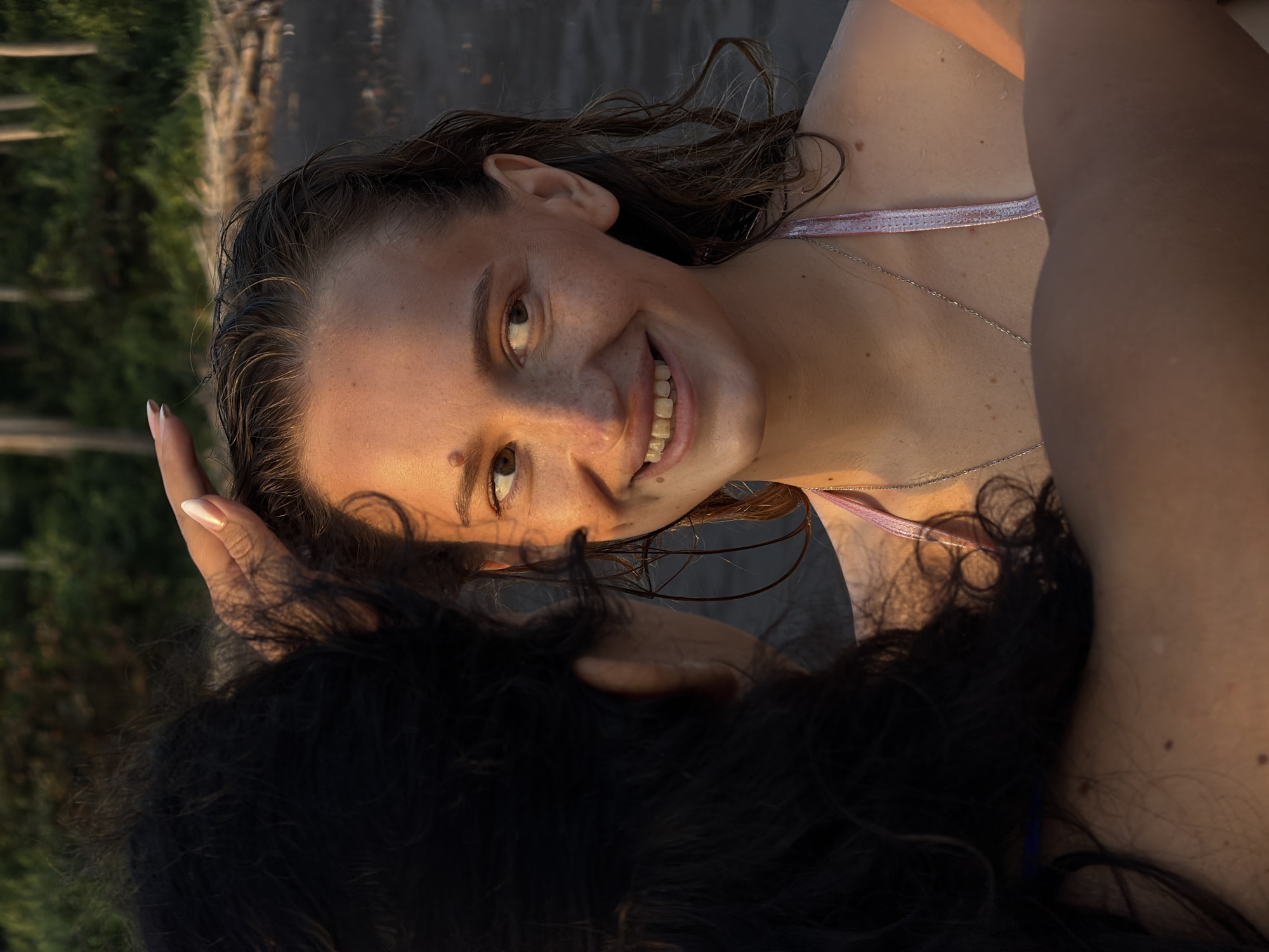 The image shows a close-up, rotated view of two people. The person in the foreground, with dark, curly hair, is partially obscuring the face of the person in the background. The person in the background, a young woman with light brown hair, is smiling and looking towards the camera. She has freckles and is wearing a bikini top. The lighting suggests it is either early morning or late afternoon, with a warm, golden hue. The background is blurred, but appears to be an outdoor setting, possibly near water, with hints of vegetation and a wooden structure. The overall mood is relaxed and intimate. No visible text is present in the image. The context provided indicates the location is Selemadeg, Indonesia.