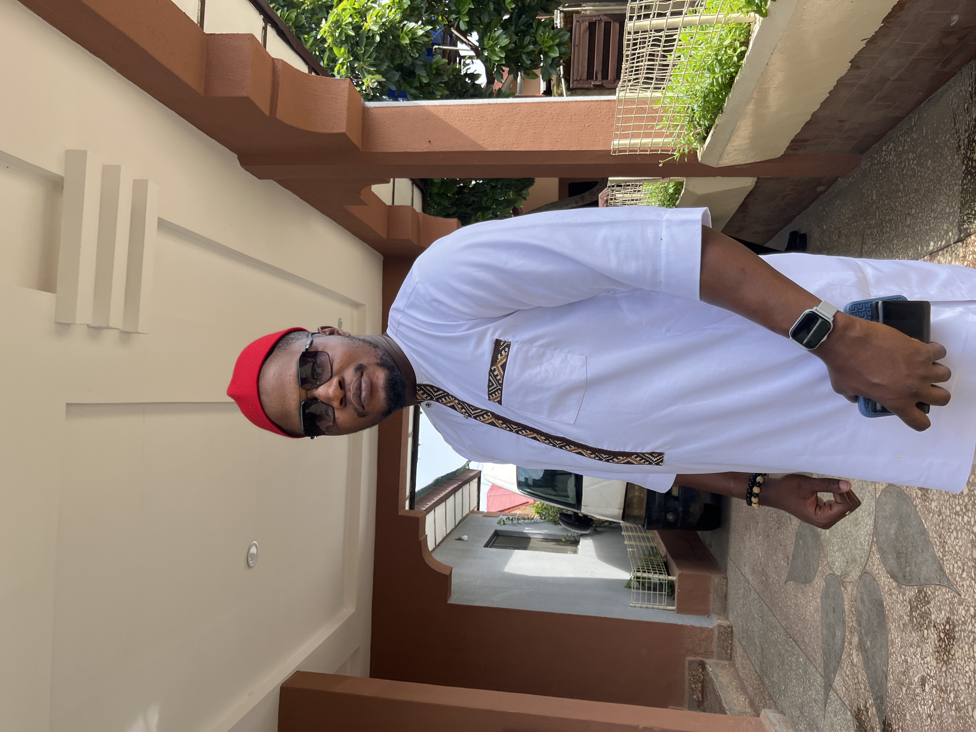 A man in his late 30s to early 40s, with dark skin and a beard, stands outdoors, facing the camera. He is wearing a white kaftan with a patterned trim on the collar and pocket, a red cap, and dark sunglasses. On his left wrist, he wears a silver smartwatch and a beaded bracelet. He holds a dark blue smartphone in his right hand. The man is standing in a covered walkway with a tiled floor. To his left, a white van is parked, and further back, a building with a grey wall and a window is visible. To his right, there is a wall with planters filled with greenery, and a metal fence. The architecture suggests a residential or commercial building. The lighting indicates it is daytime. This scene is consistent with the environment found in cities like Jos, Nigeria, characterized by this type of building design and outdoor spaces.