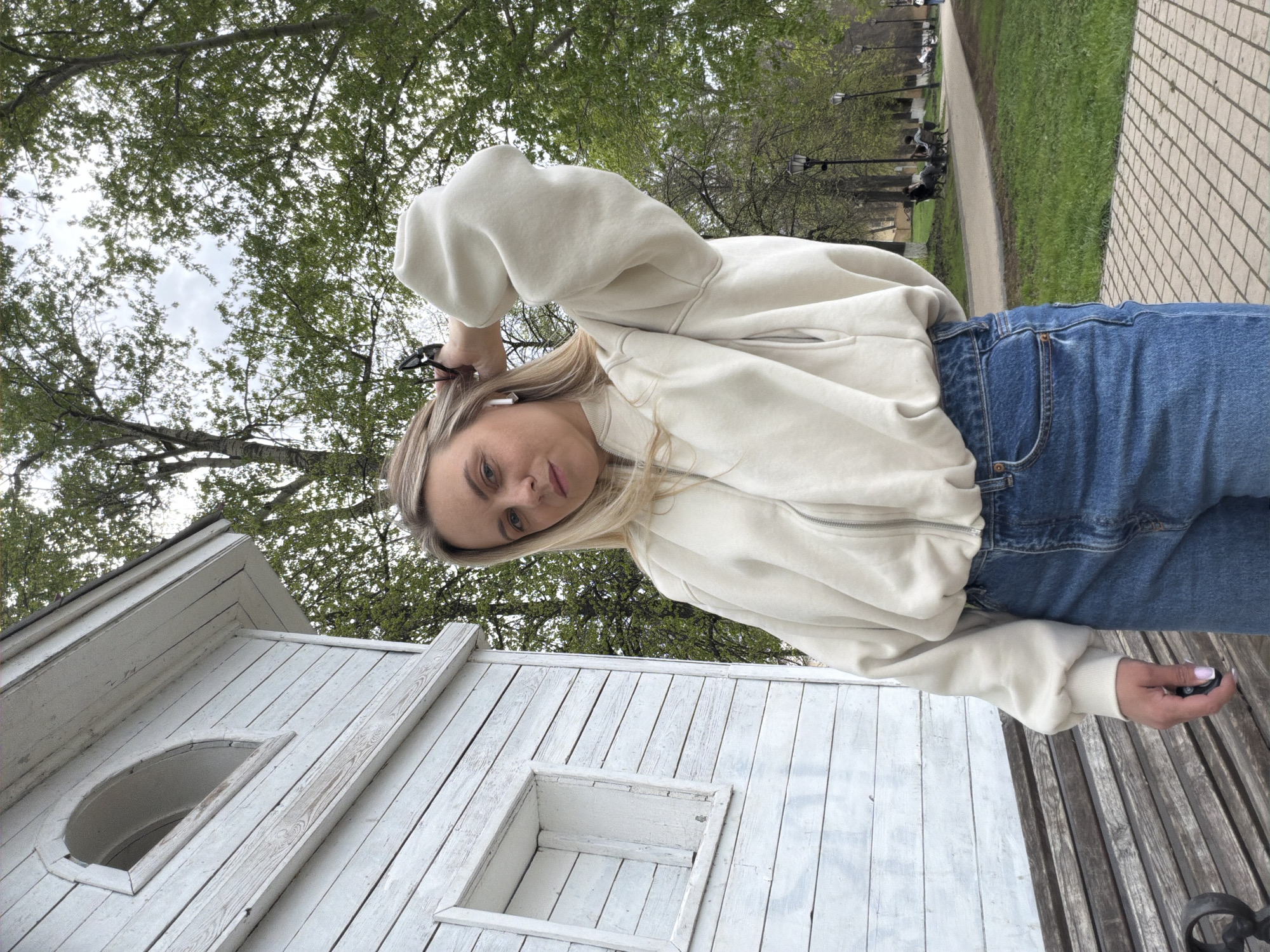 A woman is outdoors in Votkinsk, Russia. She wears a light-colored zip-up sweater and blue jeans. She holds a pair of eyeglasses in her hand and appears to be using wireless earbuds. She is near a small, white wooden structure, possibly a decorative birdhouse or small building. Behind her, there are trees and a walkway. Other people sit on benches in the background. The ground is paved with stone.